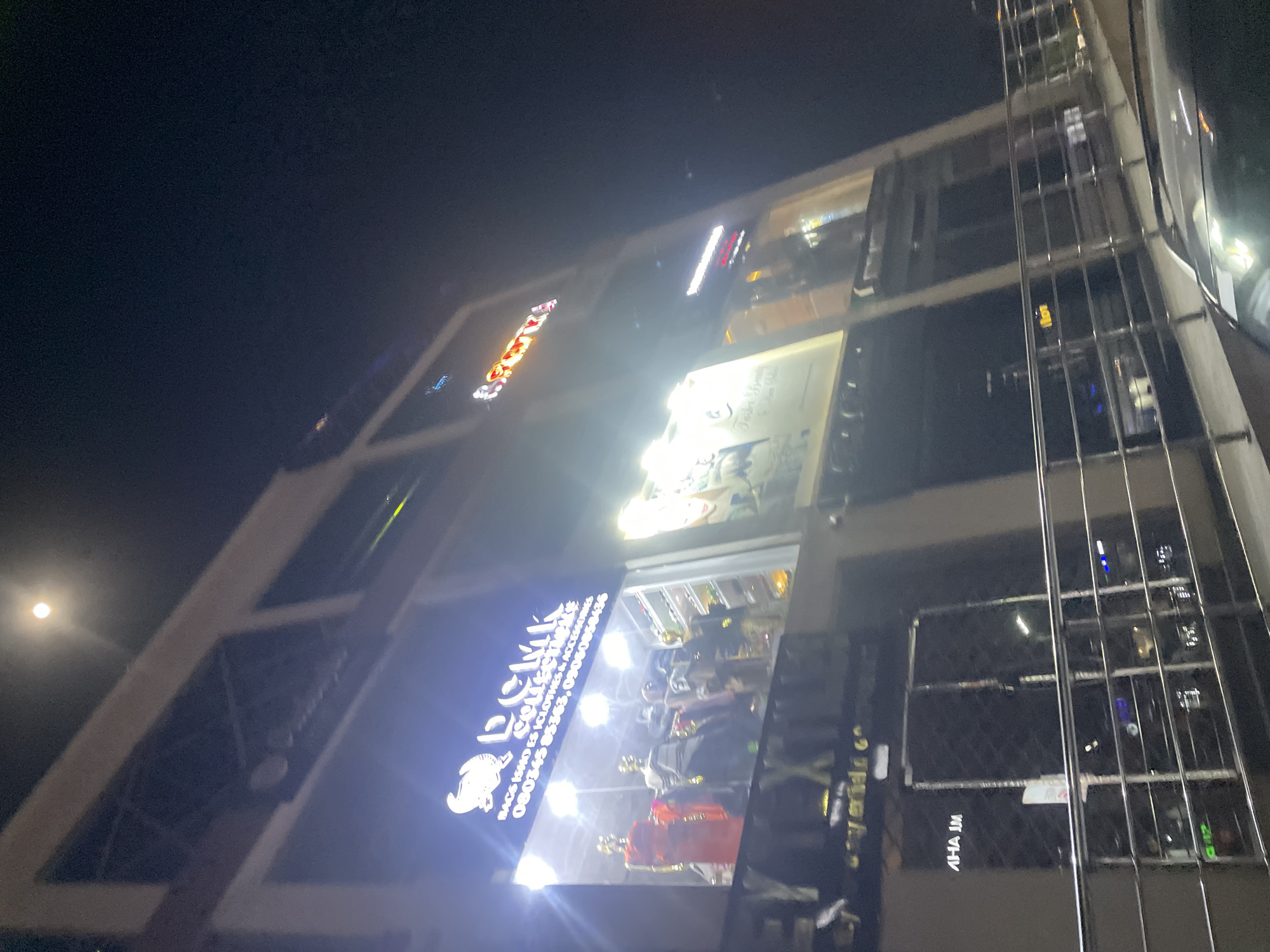At night, a multi-story building is seen in Abuja, Nigeria. Illuminated signage for "D'Clique Collectibles" is prominently displayed above a storefront window showcasing mannequins. Other signs are present on higher floors but are less distinct due to glare. The sky is dark with a bright, full moon. The building's architecture features a modern design with visible structural supports on one side.