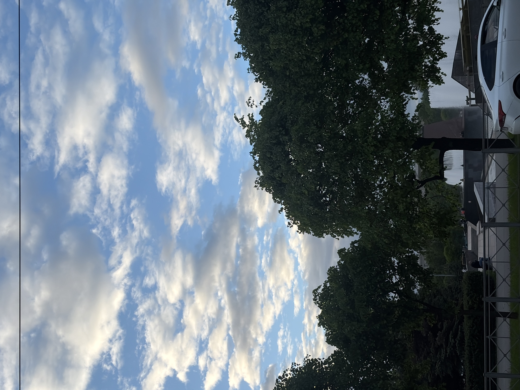 The image shows a view of Saint Petersburg, Russia, with a blue sky featuring scattered cumulus clouds partially obscured by tree foliage on the right. A white car is visible near a fence, with glimpses of buildings beyond the trees. A person is sitting on a bench in the distance.
