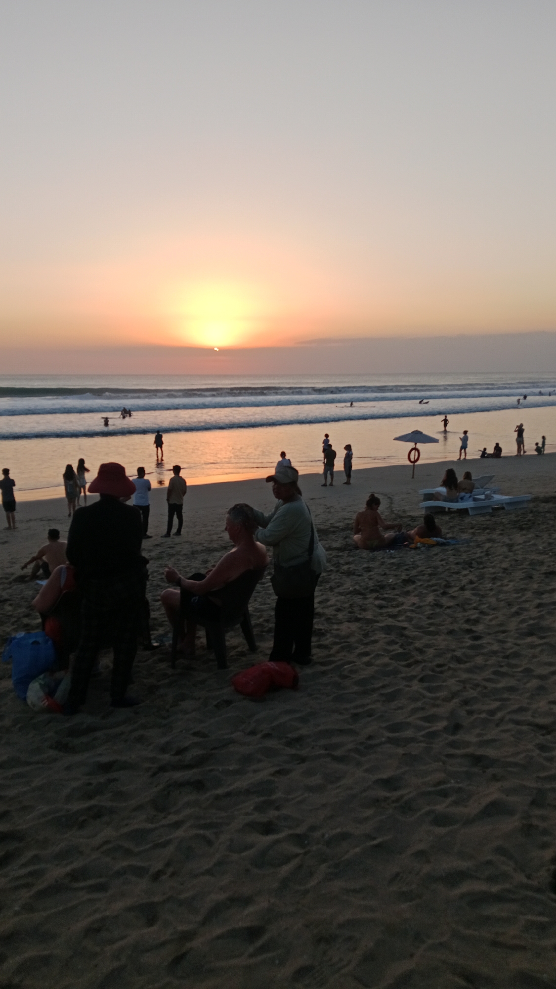 The image depicts a sunset scene at Kuta Beach, Bali, Indonesia, with numerous people present on the sandy shore and in the ocean. The sun is low on the horizon, casting a warm orange and yellow glow across the sky and water, with gentle waves breaking on the beach.
In the foreground, a person wearing a red hat and dark clothing stands near another person seated in a dark chair. A third person, wearing a light-colored shirt and a bag over their shoulder, appears to be attending to the seated individual, possibly giving a massage. To the left and right of these central figures are other people scattered on the sand, some sitting, some standing.
Further back, closer to the water, a group of people are visible. Some are standing, some are seated on loungers, and a couple are interacting near an umbrella with a life preserver. Several individuals are wading or swimming in the ocean, and a few are seen on surfboards.
The overall atmosphere is relaxed, with beachgoers enjoying the end of the day and the ocean views. The sand is textured with ripples from the wind and foot traffic. Various items, including bags and a red cushion, are present on the sand.
