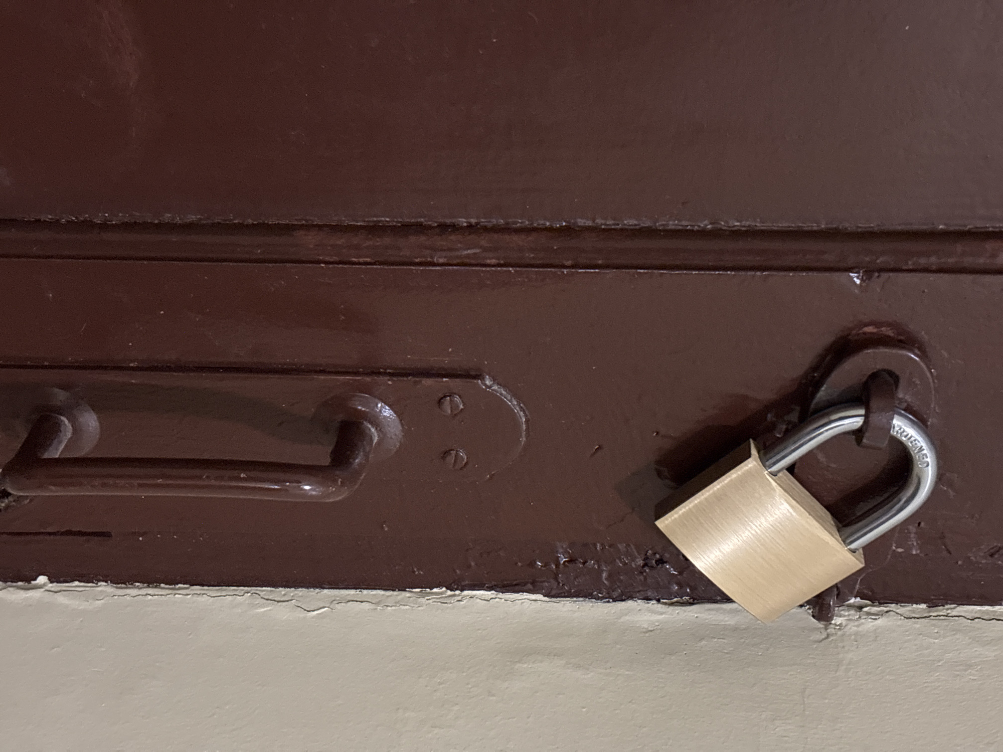 A close-up, eye-level shot shows a brass padlock secured to a dark brown wooden door, possibly an interior door in an apartment building in Tallinn, Estonia. The padlock's shackle is partially obscured by the door frame, and the body of the padlock rests against the frame. To the right of the door, a light beige wall with a cracked paint finish is visible, suggesting wear and tear in the environment. Below the padlock, a dark metal door handle is mounted on the door. The overall impression is one of a functional but aged entryway.