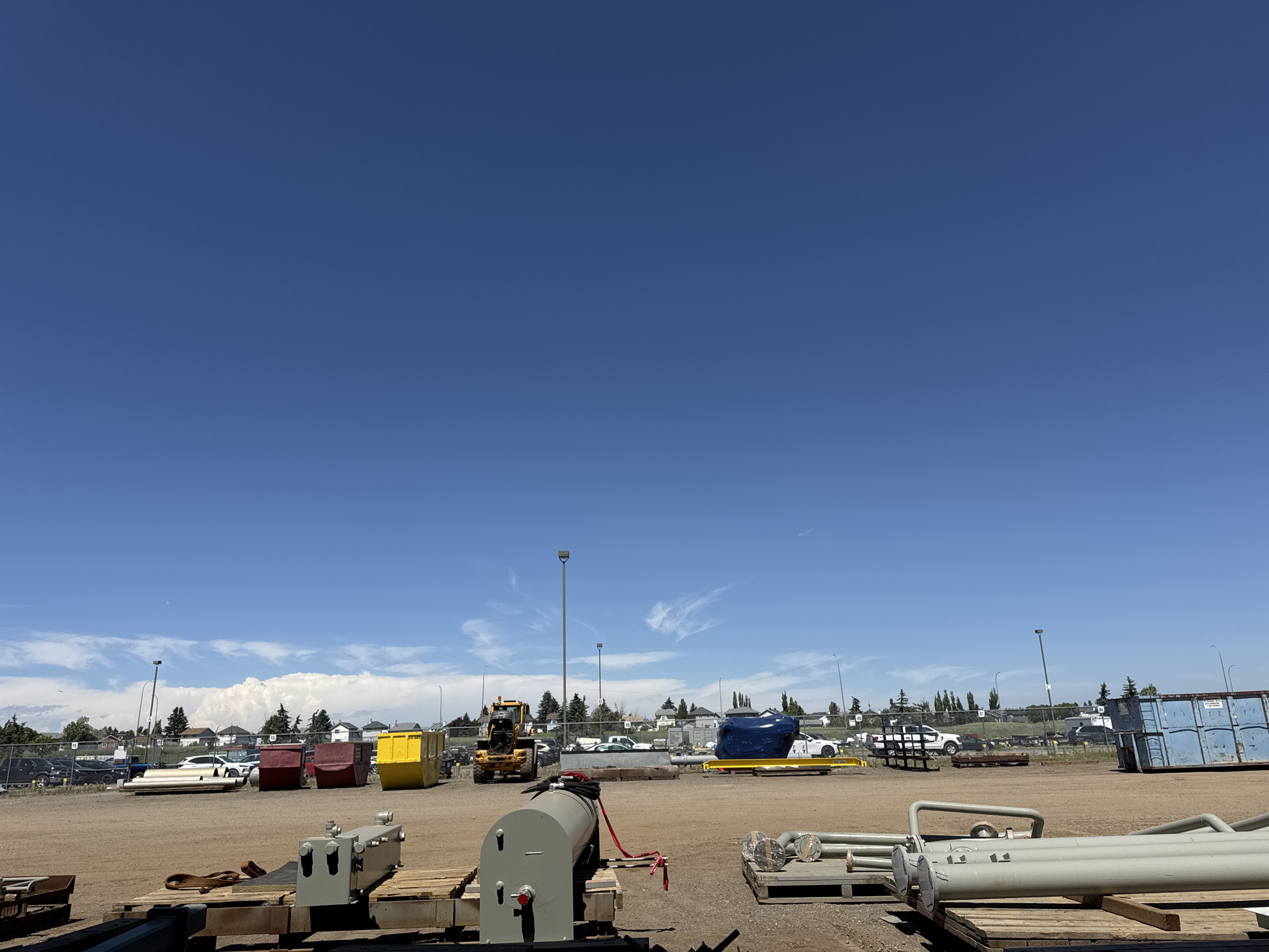 This is a wide, low-angle, outdoor, daytime shot of a storage yard with a clear blue sky. The yard is filled with construction materials and equipment. In the foreground, there are several large metal pipes and industrial machinery. In the midground, there are various colored dumpsters, including red, yellow, and blue, as well as a yellow backhoe loader and a blue tarped object. In the background, there is a residential area with houses and trees, and a chain-link fence. Streetlights are visible throughout the yard. The overall impression is one of an industrial or construction site. The scene is located in Calgary, Canada.