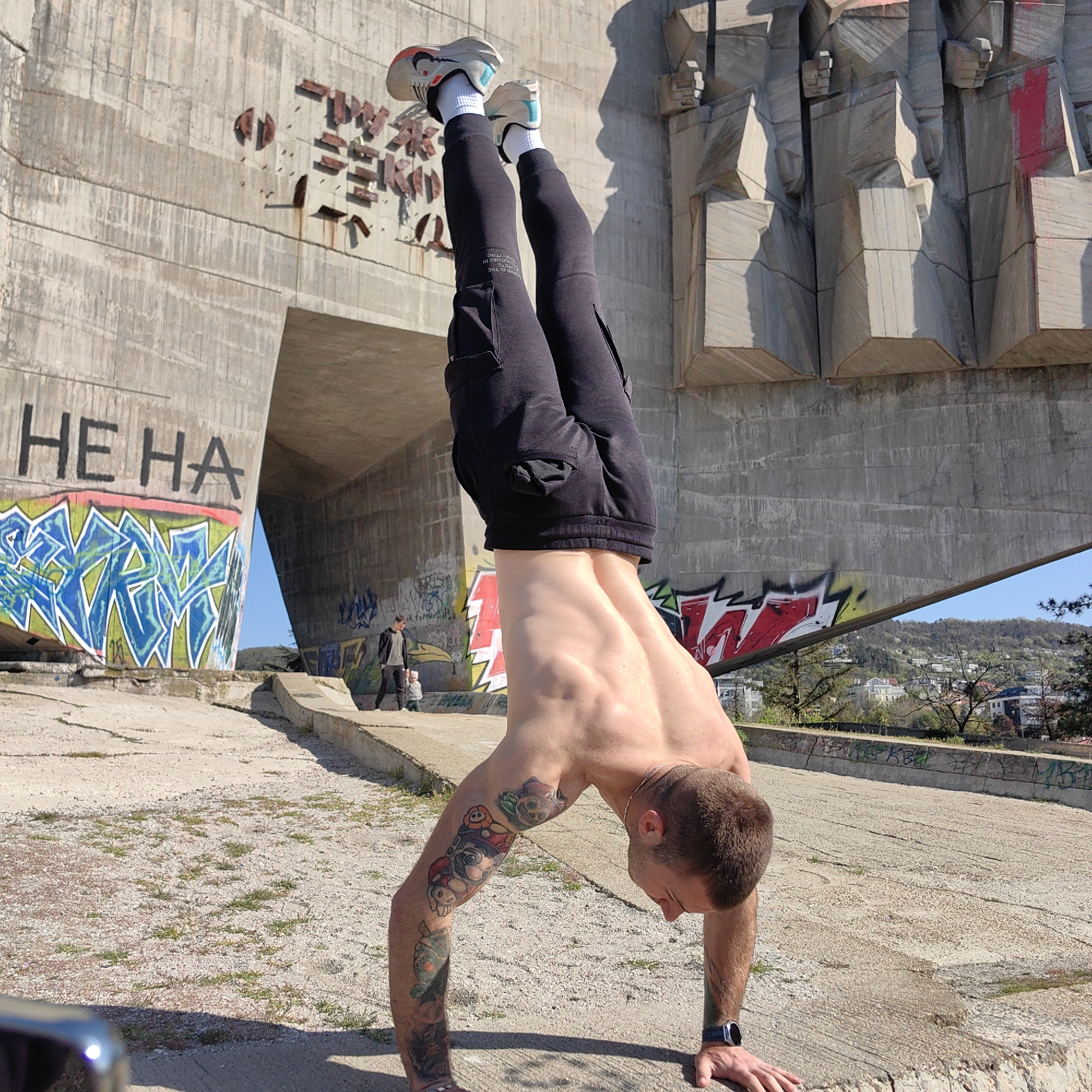The image features a muscular, bare-chested man performing a handstand on a rough concrete surface, demonstrating strength and balance. He wears black athletic joggers with text that appears to read "PROJECTED DESIGN" on the left leg, white socks, and light-colored athletic shoes with orange and teal accents. His left arm is adorned with colorful tattoos, including cartoon characters. He has short brown hair and a smartwatch on his left wrist, and his face shows concentration.
The setting is a large, imposing concrete structure, indicative of Brutalist architecture, likely a monument or bridge in Varna, Bulgaria. On the left, a massive concrete wall features black block letters spelling "HE HA" above a vibrant blue, green, and white graffiti tag. The structure's right side is composed of abstract concrete sculptural elements, possibly depicting figures, some stained with red spray paint and showing weathered metal fragments. The ground is a mix of cracked concrete and gravel with patches of green grass. In the background, through an opening in the structure, two blurry figures, an adult and a child, are visible. Beyond them, a hillside dotted with trees and buildings suggests an urban landscape. The bright sunlight casts strong shadows, indicating a clear, sunny day.