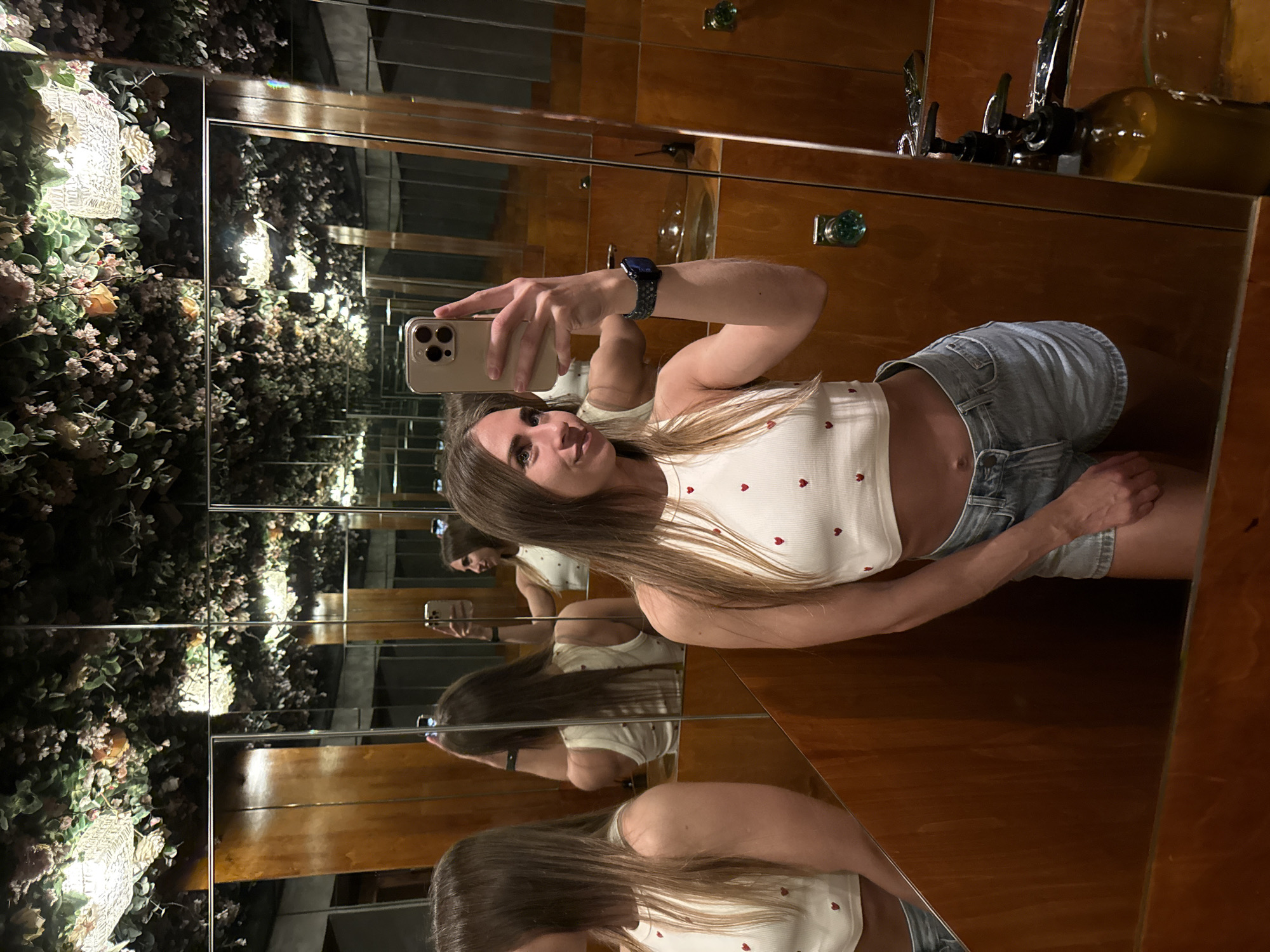 The media file shows a young woman taking a selfie in a mirrored room. She is wearing a white crop top with small red hearts and light-wash denim shorts. Her left arm is extended, holding a smartphone to capture the image. She has long, light brown hair and is looking at the phone screen with a slight smile.
The setting appears to be a bathroom or dressing area with mirrored walls and wooden paneling. The mirrors create a series of reflections of the woman and the surrounding environment, giving the impression of an infinite space. In the background, reflected in the mirrors, are elements of decor that include lush green plants, possibly artificial, and a decorative light fixture. There are also reflections of what looks like a faucet and a bottle.
The overall mood is casual and relaxed. The lighting is dim, suggesting it might be evening or indoors with controlled lighting. The reflections create a dynamic and somewhat disorienting visual effect. No visible text is present in the image.