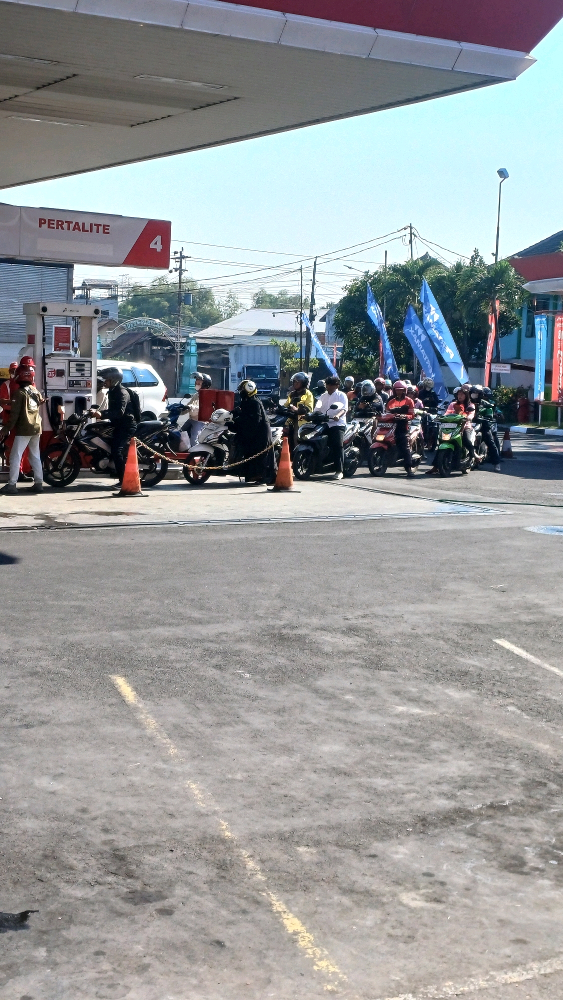 A group of people on motorcycles are waiting in line at a Pertamina gas station in Pabelan, Indonesia. The scene is outdoors, with a clear blue sky and bright sunlight. The gas station has a red and white awning, and a prominent sign reading "PERTALITE" with the number "4". Several orange traffic cones are placed on the ground. The people are wearing helmets and casual clothing. Some are standing next to their motorcycles, while others are seated. The motorcycles are parked in a line, indicating they are queuing for service. In the background, there are buildings, trees, and power lines, typical of a semi-urban or suburban environment. Blue flags with white lettering are visible behind the line of motorcycles, suggesting a possible promotional event or organized group activity. The ground in front of the gas station is asphalt, marked with yellow lines.