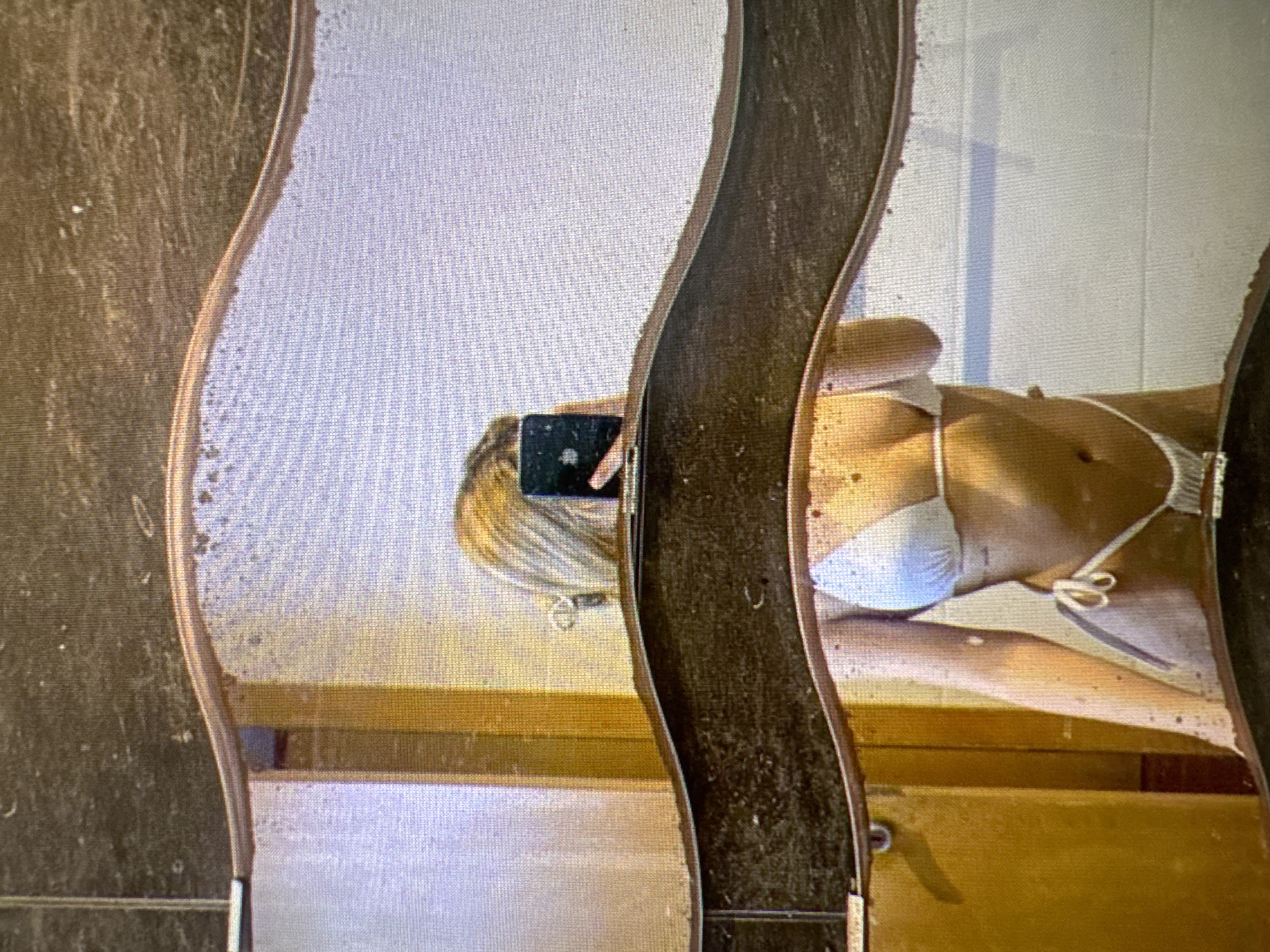 The image shows a reflection in a wavy mirror. A person wearing a white bikini is visible in the reflection, holding a phone to take the picture. The background appears to be a bathroom with tiled walls and a wooden door. There is no information in the image to suggest the location is Castelo Branco, Portugal.