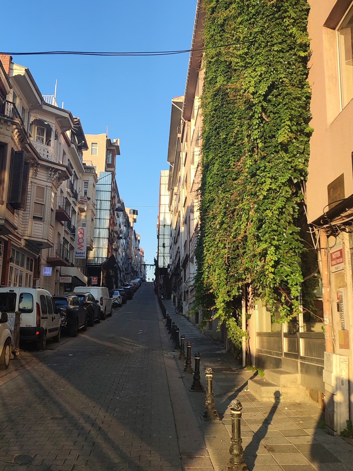 The image shows a narrow, uphill street in Kadıköy, Turkey, lined with buildings of varying architectural styles. The street is paved with cobblestones and is partially shaded by the buildings and the bright sunlight casting long shadows. Cars are parked along both sides of the street, including white vans and darker sedans. On the left side of the street, buildings feature traditional Ottoman architecture with ornate balconies and bay windows. Further up the street, modern buildings with glass facades are visible. On the right side, a building is covered in dense green ivy, with a pale pink facade. The sky is clear and blue. In the distance, at the top of the hill, two figures can be seen walking. The overall impression is of a charming and historic urban street scene in the afternoon light.
