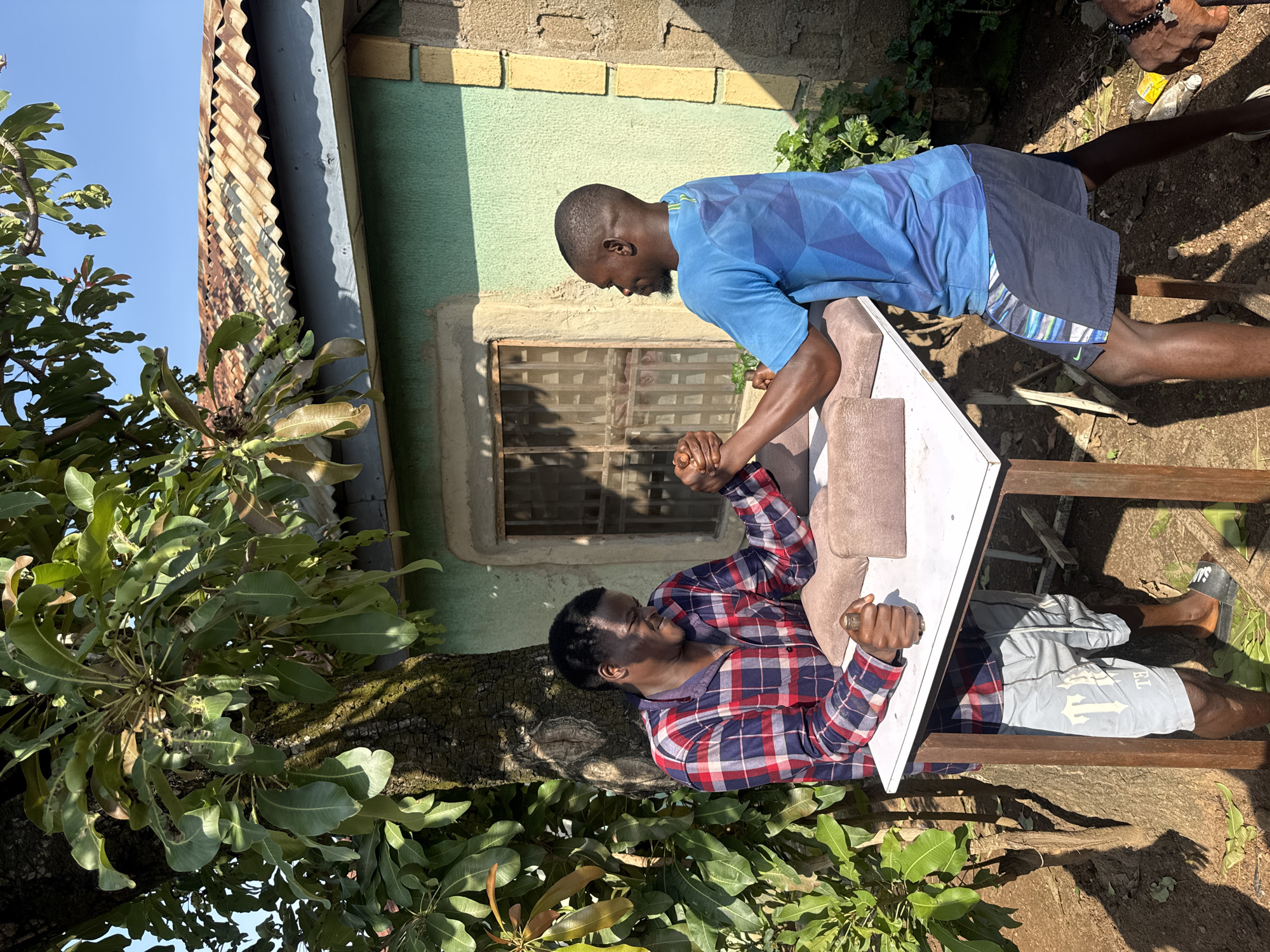 Two young Black men are arm wrestling at a table outdoors in Akwanga, Nigeria. The man on the left, wearing a red and blue plaid shirt and grey shorts, strains with his arm on a padded rest. The man on the right, wearing a blue t-shirt and dark shorts, grips his opponent's hand and appears to be winning. They are positioned at a white table with brown padding, under the shade of a large tree with green leaves. Behind them is a pale green building with a corrugated metal roof and a barred window. The ground is dirt, with some fallen leaves and sparse vegetation. The lighting suggests it is daytime.