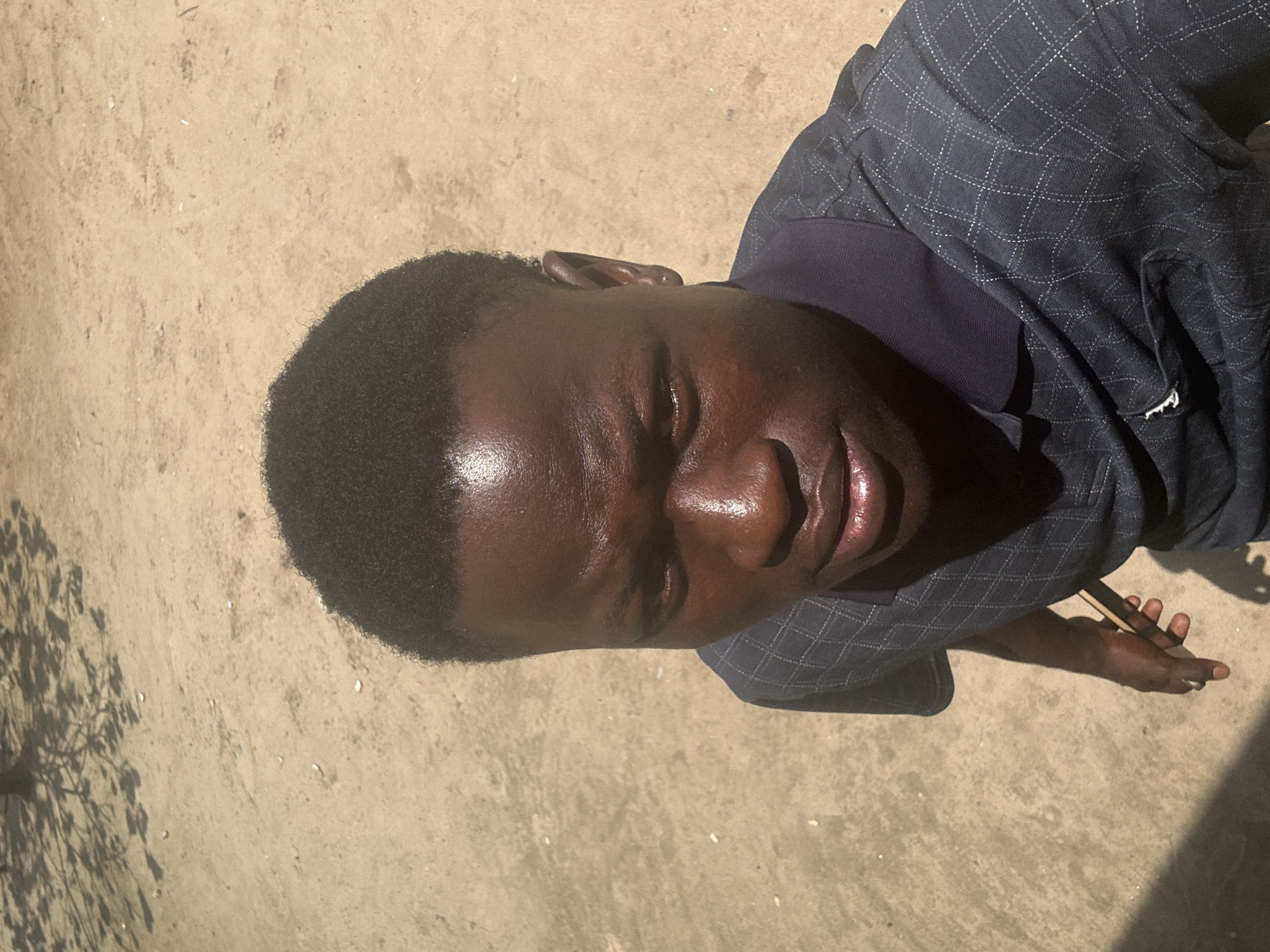 A high-angle medium shot captures a young Black male from the chest up, standing outdoors in what appears to be Ganyingbo, Nigeria. He is facing directly towards the camera with a neutral expression. The background is a dusty, sandy ground with dappled sunlight and shadows cast from unseen foliage overhead. He wears a dark blue collared polo shirt with a subtle diamond pattern. His skin is dark and shiny, suggesting sweat or oil. His hair is short and dark, styled in a close-cropped cut. In his right hand, he holds a thin, light-colored object, possibly a stick or a cigarette, between his thumb and forefinger. The overall scene suggests a casual, outdoor environment, possibly during daylight hours given the strong shadows.