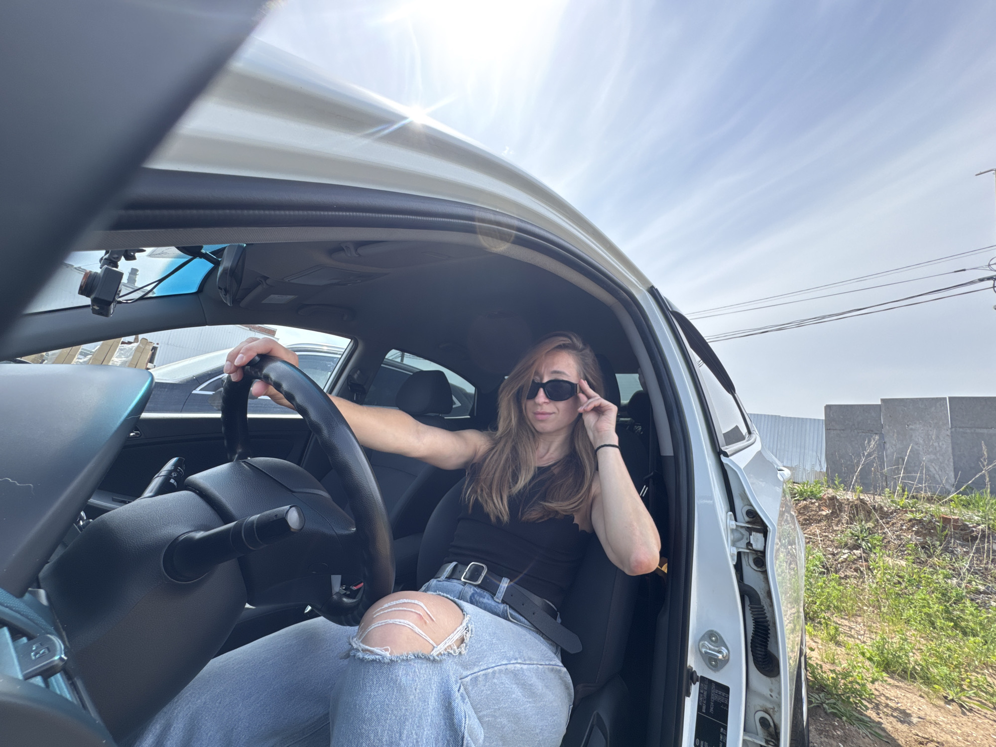 A woman is seated in the driver's seat of a white car. She is wearing sunglasses, a black tank top, and ripped blue jeans with a black belt. Her right hand is on her sunglasses and her left hand is on the steering wheel. The car is parked on a sunny day. Concrete blocks and a metal fence are visible outside of the car. The location is Chaikovsky, Russia.