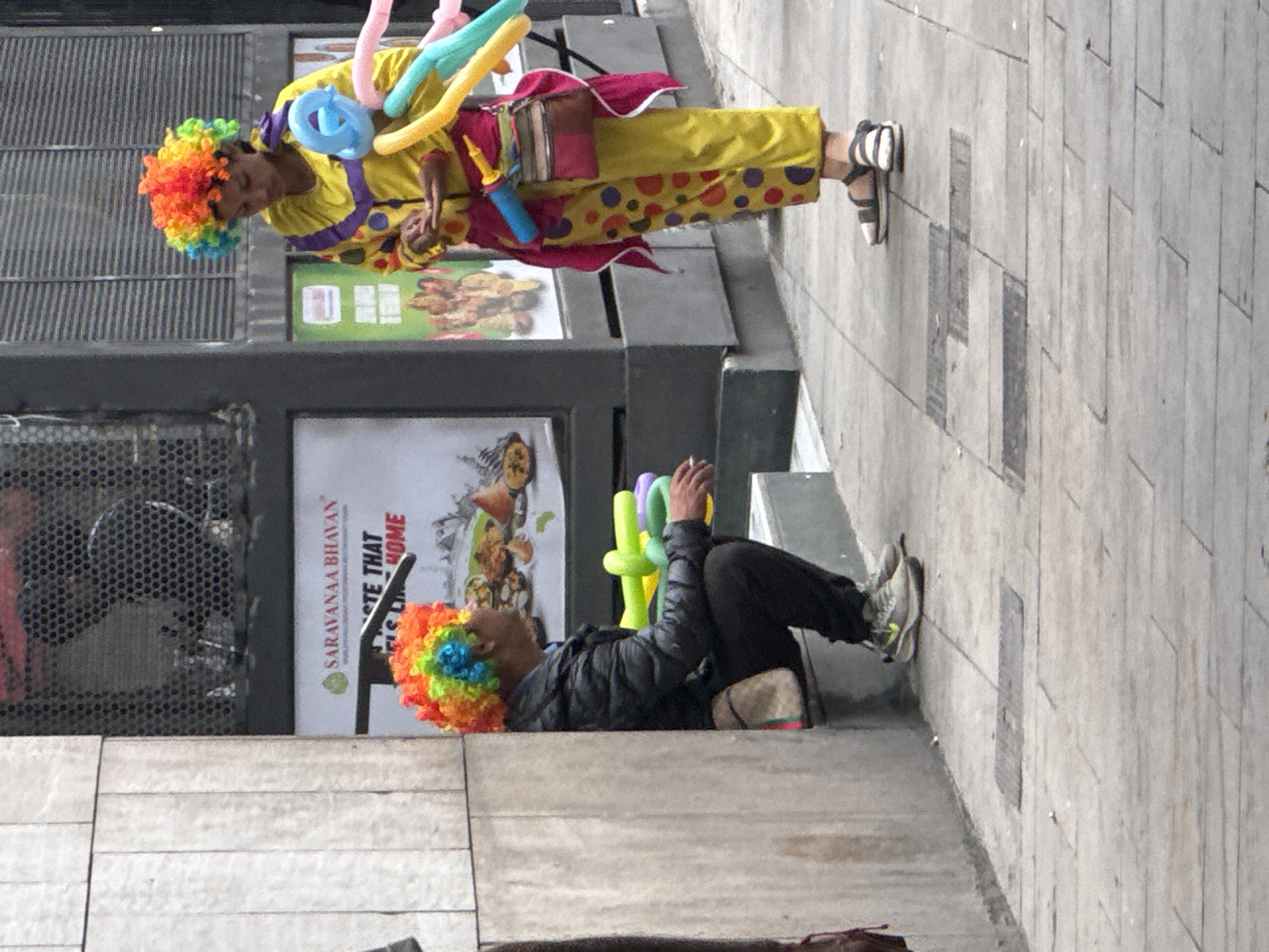 Two people in clown wigs are seen in this image. One is standing in a yellow and red polka-dotted outfit, holding balloons. The other is lying on the ground making balloon animals. The scene is set in an urban area, with a sign that reads "Saravanaa Bhavan Taste that Like Home". Based on the sign, it is likely located in Barcelona, Spain.