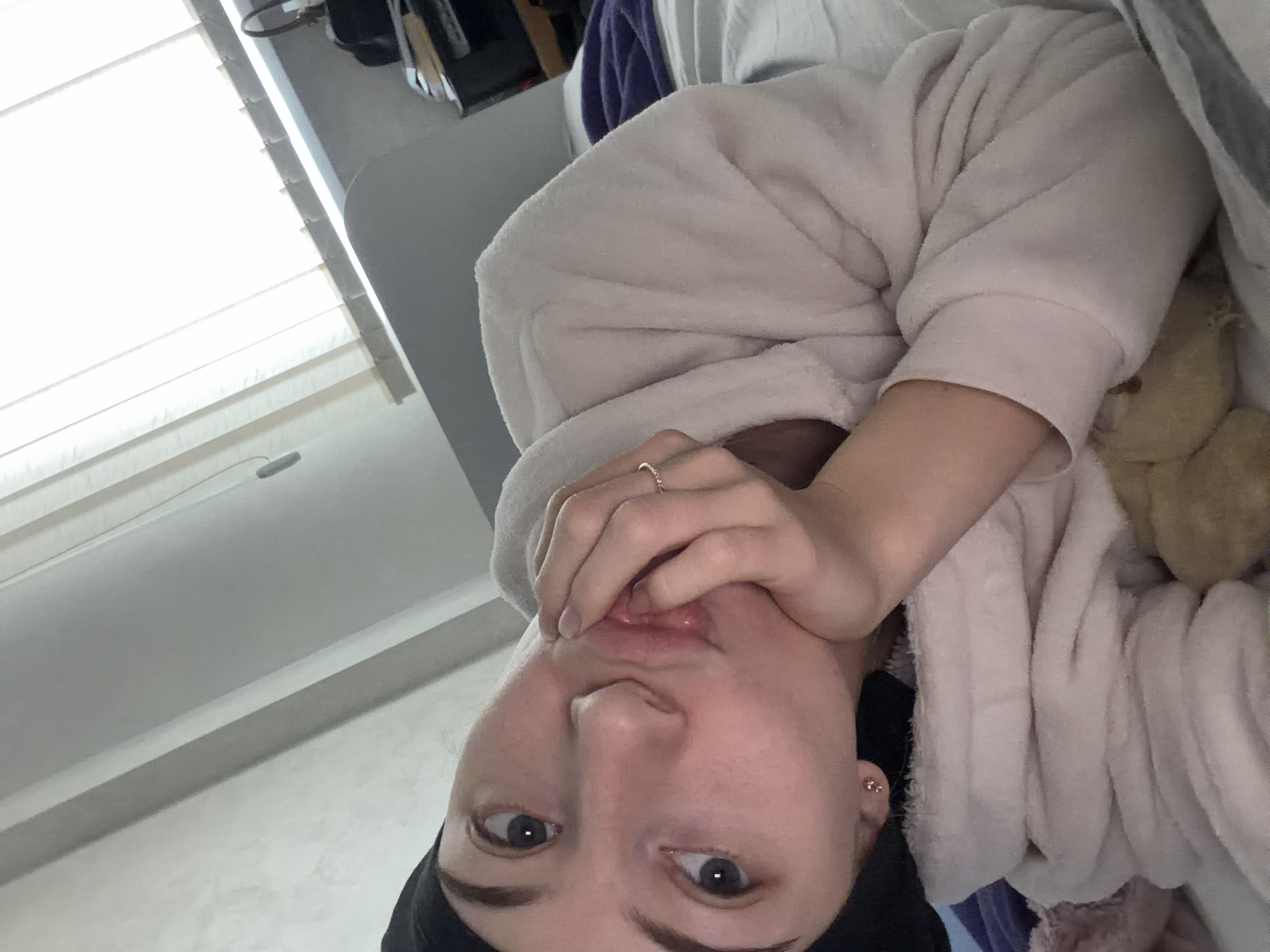 A selfie of a person lying in bed in a room with blinds covering a window. The person is fair-skinned, wearing a black head covering and a pink robe. They are holding their hand up to their mouth. A ring is visible on one of their fingers. Part of a stuffed animal is visible near the person's arm. The location is unknown and cannot be determined from the image, and there is no way to confirm that this is Plovdiv, Bulgaria.