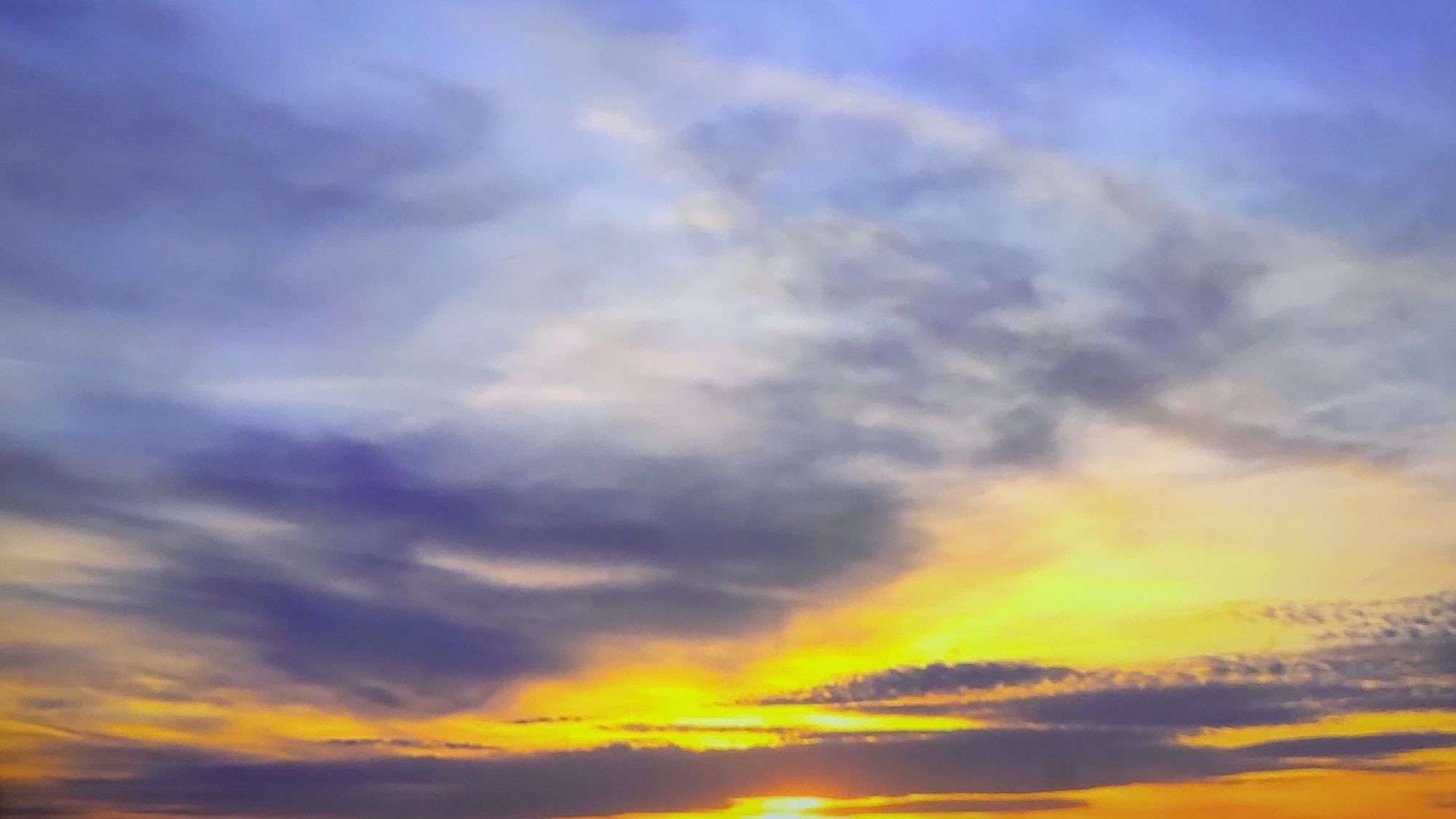 The image shows a vibrant sunset with hues of yellow, orange, and purple. The sky is filled with clouds, some appearing dark and heavy, while others are illuminated by the setting sun. There is no evidence to suggest this image was taken in Yerevan, Armenia.