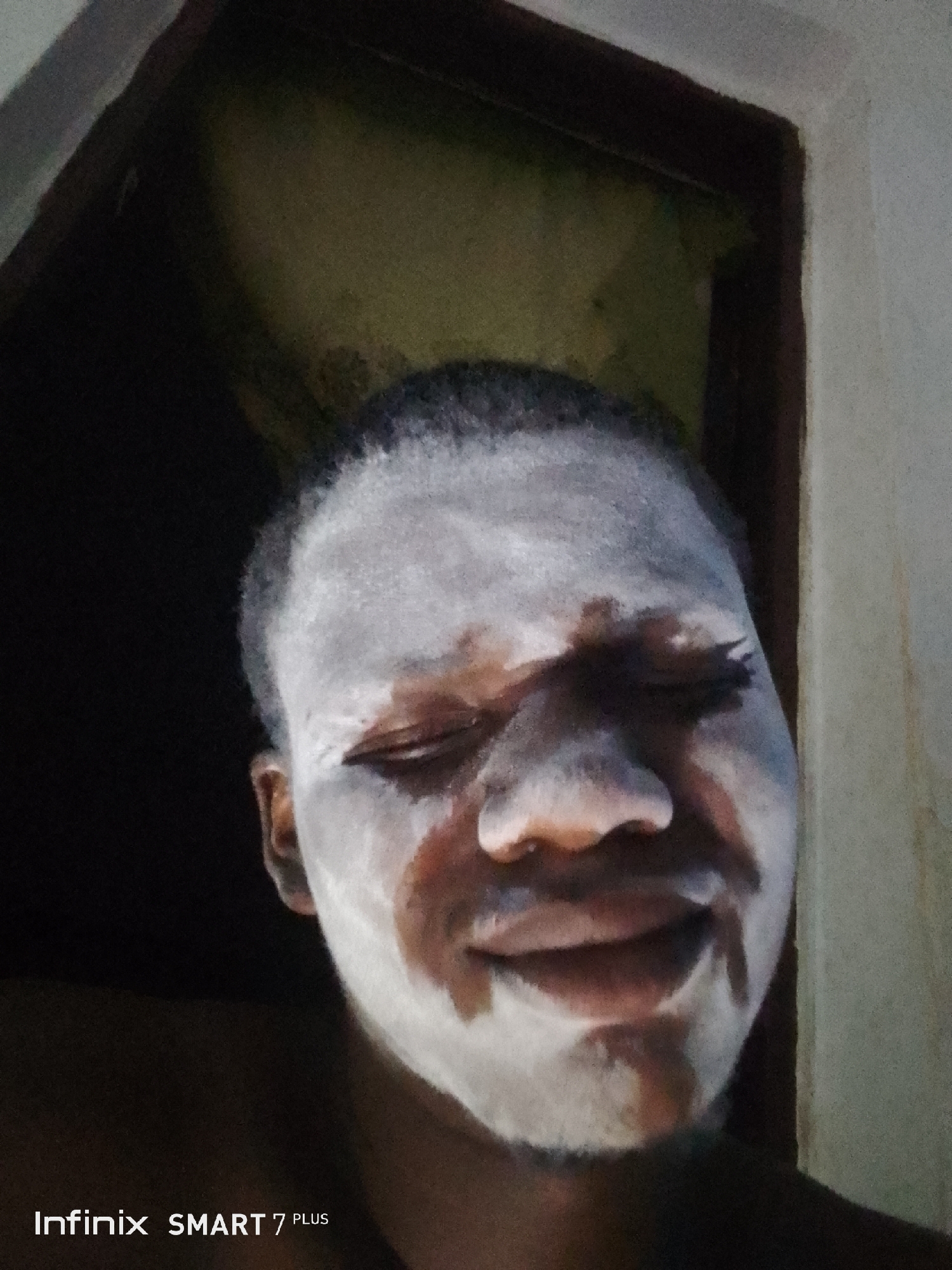 A close-up portrait shows a person with dark skin, whose face is partially covered in an unevenly applied white substance, resembling face paint or a mask. The person has short, dark hair and appears to be smiling or laughing, with their eyes closed or squinted and their mouth slightly open, conveying an emotion of amusement or joy. The white substance is prominent on their forehead, cheeks, and chin, with areas of their natural skin tone visible underneath.
The background is dark and indistinct, suggesting an indoor environment. A dark, possibly wooden, frame runs along the top left and right sides, indicating a doorway or a corner of a room. A lighter, textured surface is faintly visible on the far right. The lighting is dim, focusing on the person's face. In the bottom left corner, the text "Infinix SMART 7 PLUS" is visible, identifying the device used to capture the image.