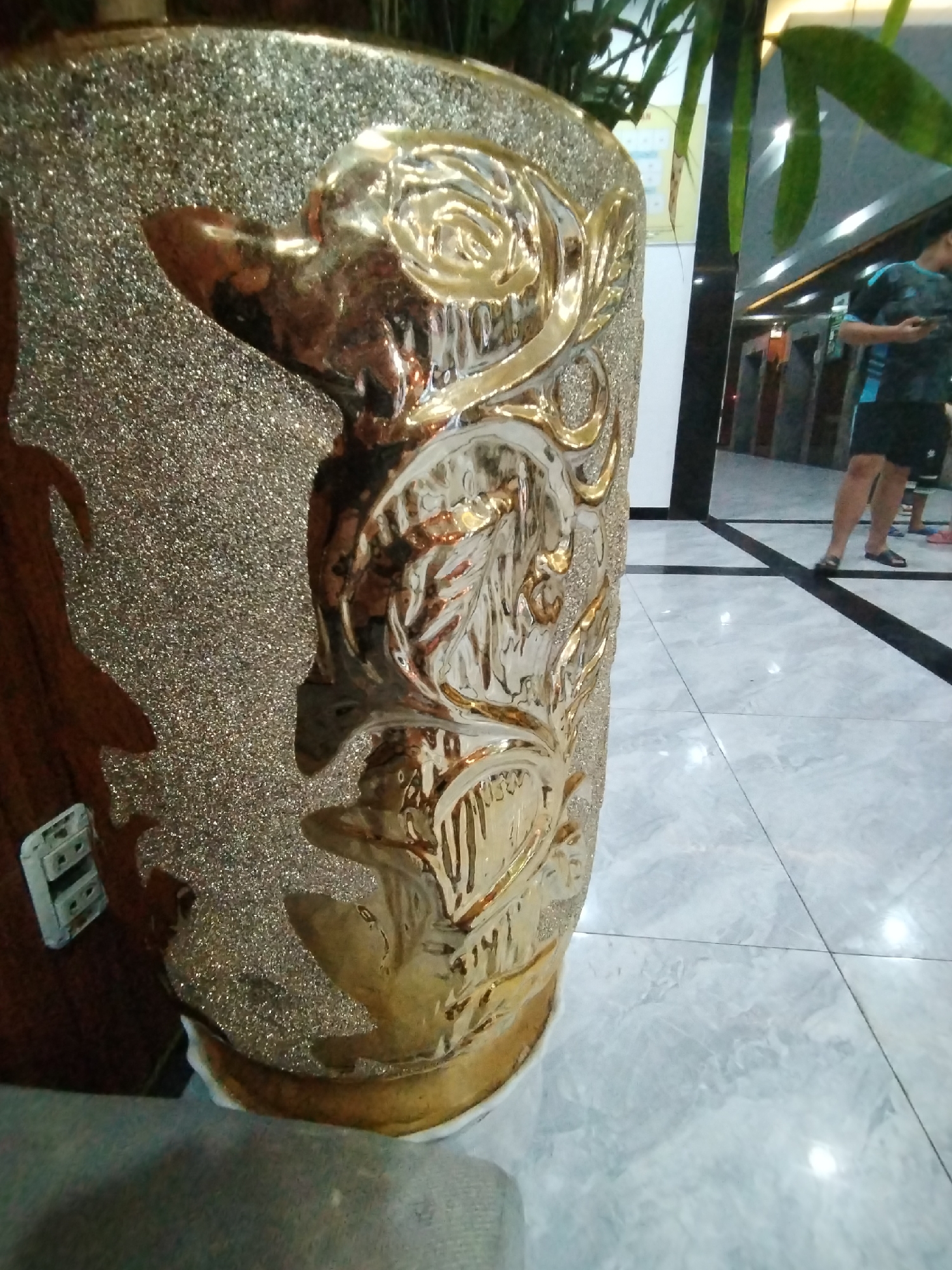 The image depicts an indoor scene, likely a modern building's hallway or lobby, located in Bac Nha Trang Ward, Vietnam. In the foreground, a large, ornate golden vase or planter dominates the left side of the frame. This decorative object features a textured, glittery golden base, adorned with highly reflective, raised golden patterns that resemble abstract swirls and stylized floral or leaf motifs. The glossy surface of these patterns reflects light from the surroundings.
From the top of the vase, green foliage, possibly bamboo, extends upwards, with some leaves visible in the upper right section of the image.
In the background, the setting includes a light-colored, possibly marble, tiled floor with dark grout lines forming a grid, which reflects ambient light. A person is partially visible in the mid-ground, out of focus. This individual is wearing a dark t-shirt with light blue accents and black shorts, and appears to be holding and looking at a smartphone. Their legs and feet, wearing dark sandals, are visible. The feet of another person are faintly discernible behind them. The background also reveals dark vertical panels and what seem to be elevator doors or architectural wall features.
The activity taking place is simply people standing or moving through the indoor space, with one person engaged with their phone. No specific event is evident. The lighting suggests an indoor, likely artificially lit, environment. There is no visible text in the image.