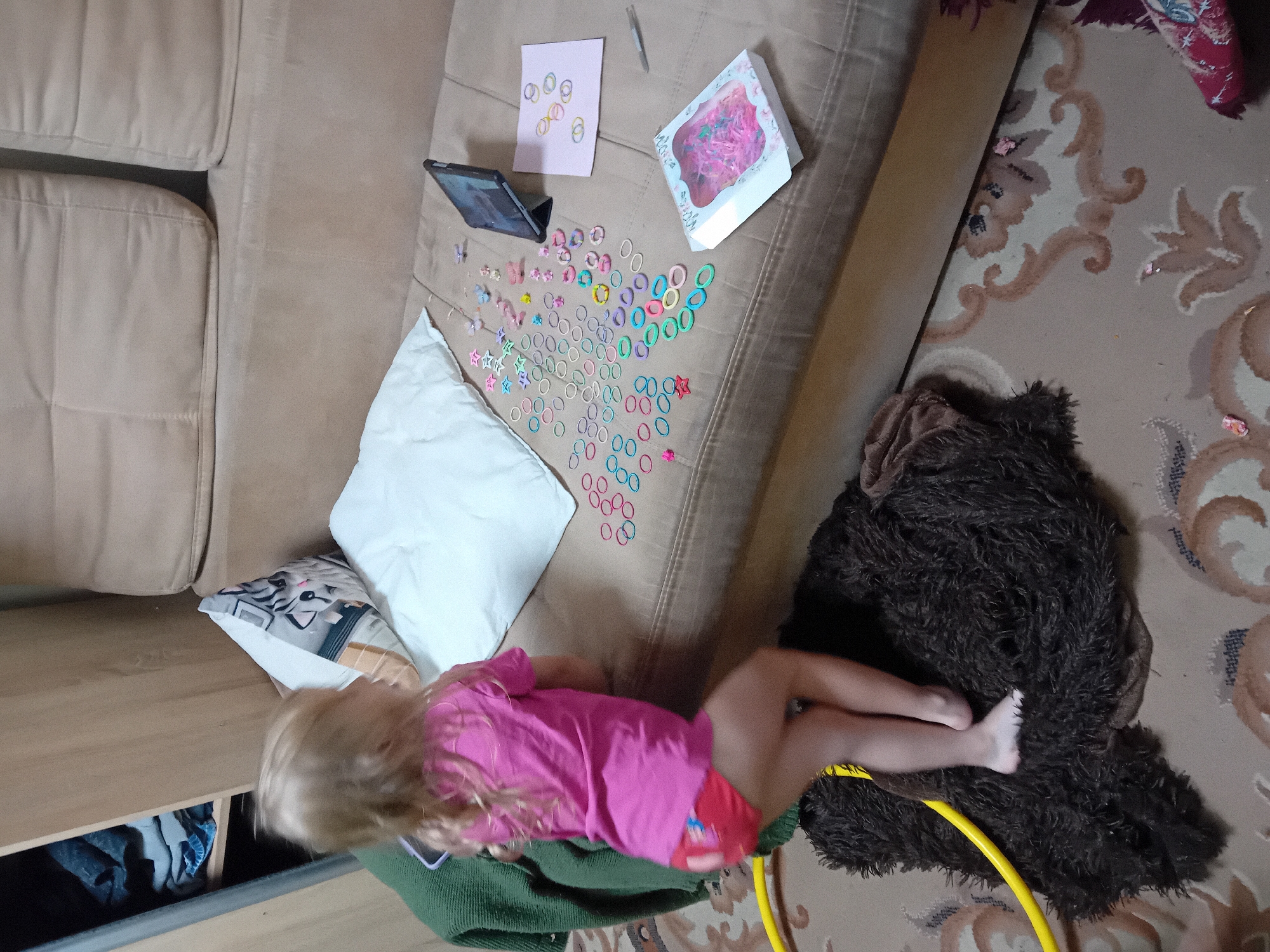 A blonde child, wearing a pink t-shirt and red shorts, stands on a dark brown rug on a patterned carpet. The child's back is to the camera, and they are looking towards a couch. The couch is upholstered in a light brown fabric and has several cushions. On the couch, there is a tablet, a small pink card with some colorful rubber bands, a box of pink and green beads, and a scattered assortment of colorful rubber bands of various shapes and sizes, including stars and rings. The background shows a wooden closet filled with clothes and a portion of the couch. The scene appears to be indoors, likely in a residential setting. The location is Chuhuiv, Ukraine.