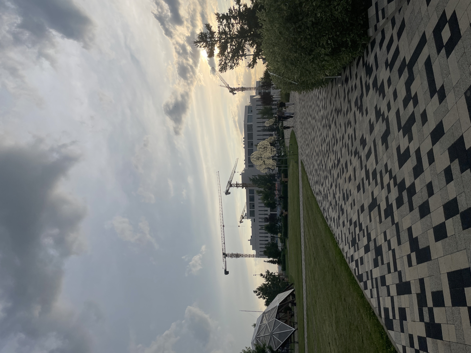 This is a medium-wide, full-shot image of a park area with a modern building and construction cranes in the background. The scene appears to be at dusk or dawn with a cloudy sky and the sun peeking through the clouds on the right side. The foreground features a patterned walkway made of light and dark rectangular stones, leading away from the viewer into the park. To the left of the walkway is a well-maintained lawn. Further into the park, on the left side, there is a geodesic dome-like structure with exposed beams and a translucent covering. Beyond this, there are several trees. In the mid-ground, a modern, multi-story building with many windows is visible, with several tall construction cranes positioned in front of and around it, indicating ongoing construction. A line of trees separates the building from the park area. To the right of the building, a tall pine tree stands in silhouette against the sky. Several people are walking along the patterned walkway, heading towards the building. The environment is urban, likely a public park within the city.