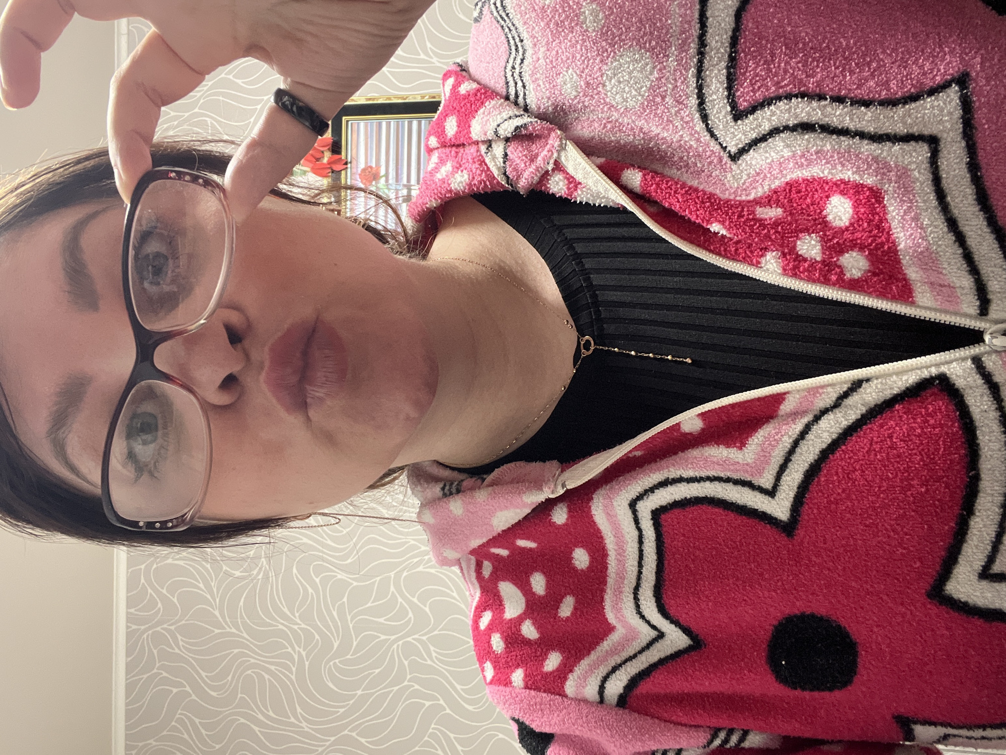 A person holds glasses in front of their face. The person is wearing a black top with a pink and white patterned jacket. The location is likely an indoor setting, suggested by the patterned wallpaper and the presence of a framed picture in the background. There is no verifiable information to confirm the city of Perm, Russia, in the scene.