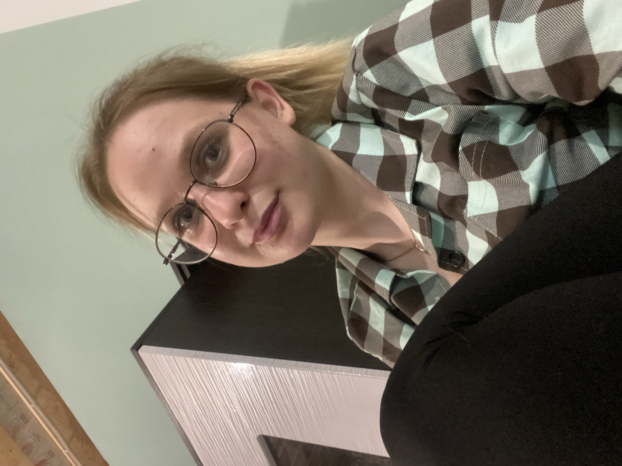 The image depicts a woman wearing glasses and a checkered shirt, likely indoors, in an unknown location. She is positioned above the camera, looking down. In the foreground, dark fabric (likely clothing) is visible, and there is a piece of furniture with a white, textured surface. The wall in the background is light green.