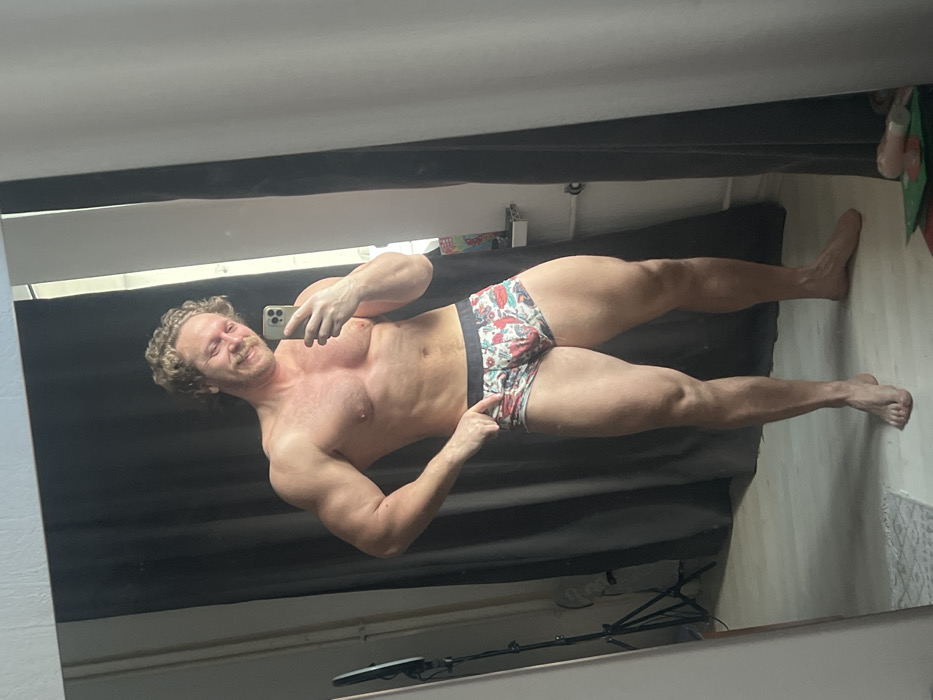 A muscular man stands in front of a mirror, posing for a selfie. He's wearing printed briefs and holding his phone with his left hand. He is fair-skinned with curly blonde hair. The background includes a dark gray curtain or backdrop and what appears to be a wooden floor, some camera equipment. His feet are visible, stretched out along the floor.
