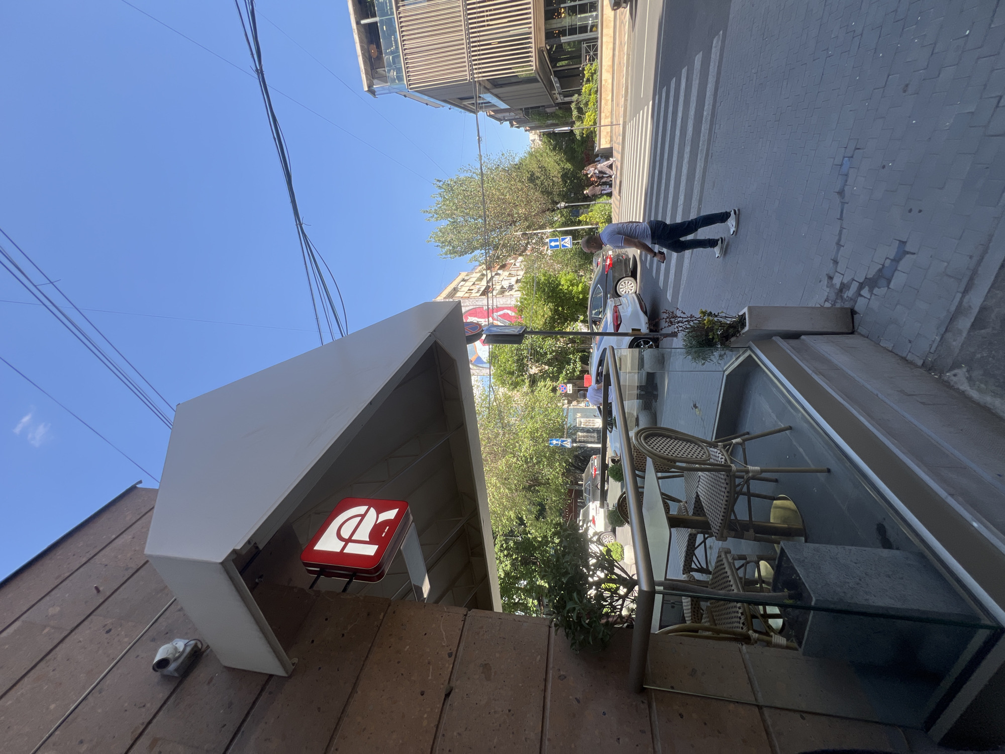 The image shows an outdoor scene in Yerevan, Armenia. A pedestrian is crossing the street at a crosswalk, near a building with a prominent red and white sign bearing a stylized Armenian letter "Պ" (P). The building appears to house a cafe or restaurant, evidenced by outdoor seating with wicker chairs and tables. A security camera is visible mounted on the building's facade. The sky is clear and blue. Other buildings are visible in the background. A parked white car is also observed next to the pedestrian.