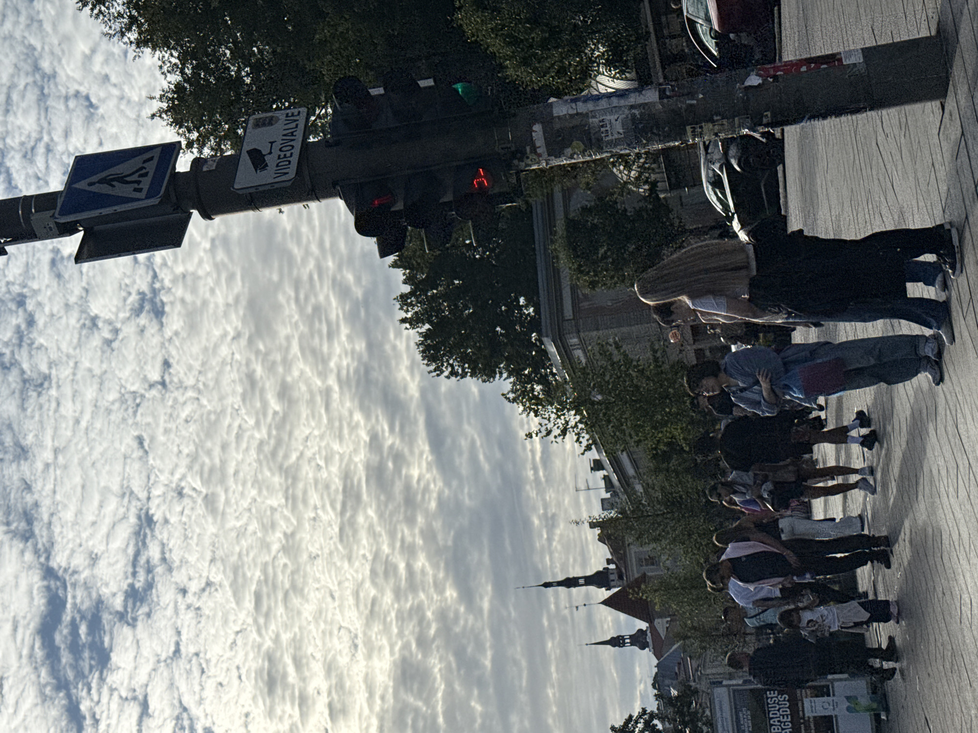The image shows a street scene in an urban environment with a traffic light pole prominently in the foreground. The traffic light has a red signal illuminated. A pedestrian crossing sign and a sign indicating "VIDEOVALVE" with a camera icon are attached to the pole. Several people are standing on the sidewalk, appearing to be waiting or walking. A young girl in a white shirt and dark pants is visible among the group. In the background, there are buildings with distinctive spires, suggesting a European city. The sky is partially cloudy with patches of blue. Cars are present on the street. The overall atmosphere appears to be daytime in a city square or intersection.
