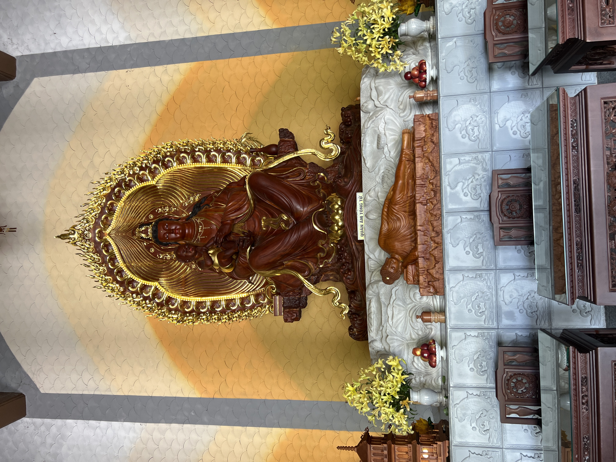 The image shows the interior of a Buddhist temple in Da Nang, Vietnam. In the center is a large, ornate wooden statue of a female deity with a baby in her lap, set against a golden, scalloped background. To the right and left of the deity, there are vases of yellow flowers. Below the deity's statue, on a white carved platform, lies a wooden statue of a reclining Buddha. In front of this platform are several red apples in a white bowl and a wooden incense holder. The foreground shows dark wooden furniture with glass tops, and ornate wooden chairs. The walls are decorated with patterned tiles. The scene depicts a religious setting with devotional objects and furniture typical of a temple.