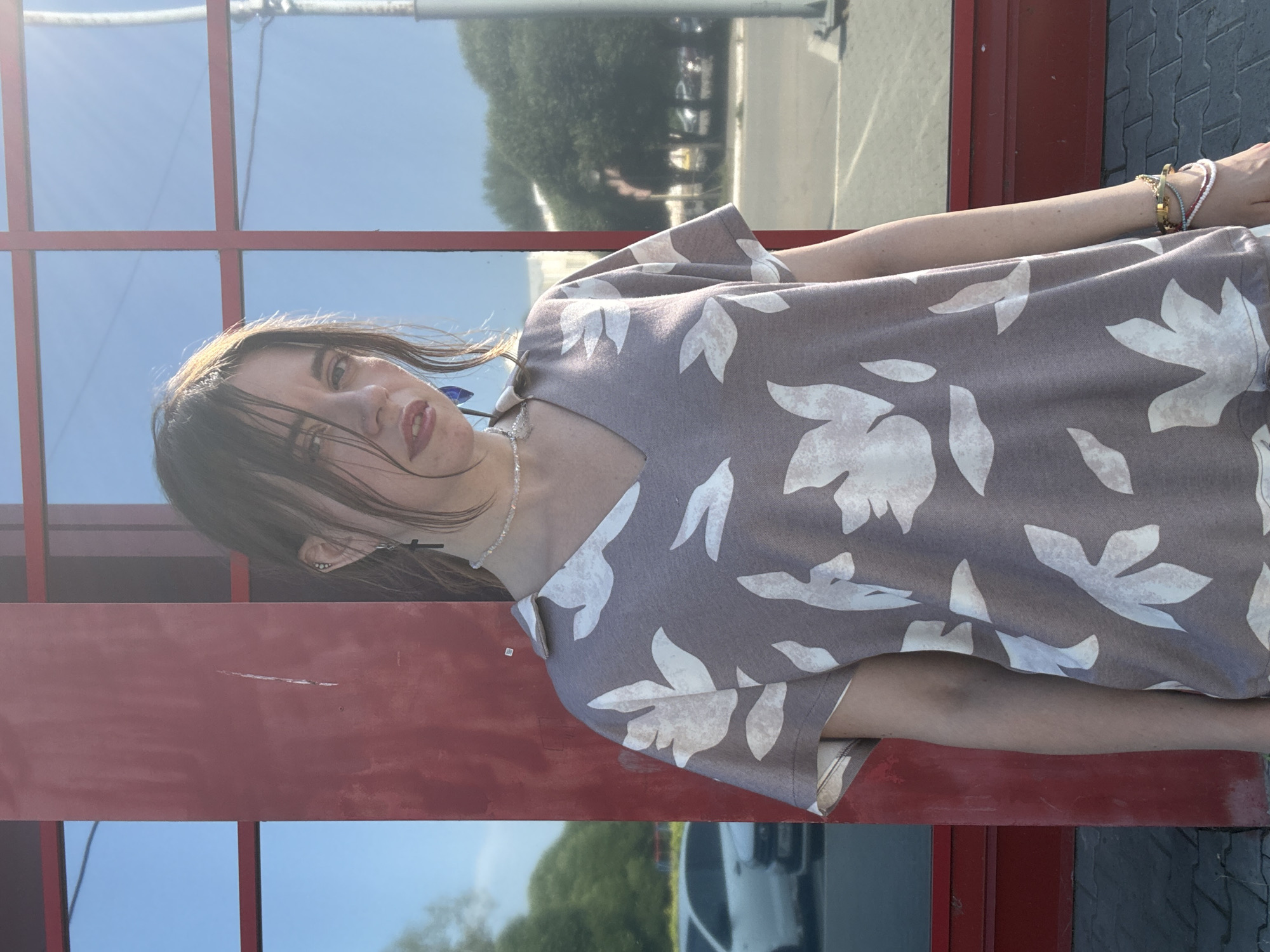 A young woman with dark hair and fair skin stands in front of a building with large red-framed windows. She is wearing a short-sleeved, V-neck shirt with a floral pattern in white on a brown background. She has on a delicate necklace and blue earrings. On her left wrist, she wears several bracelets. The background shows a reflection of trees and a street with a white car. The lighting suggests it is a sunny day.