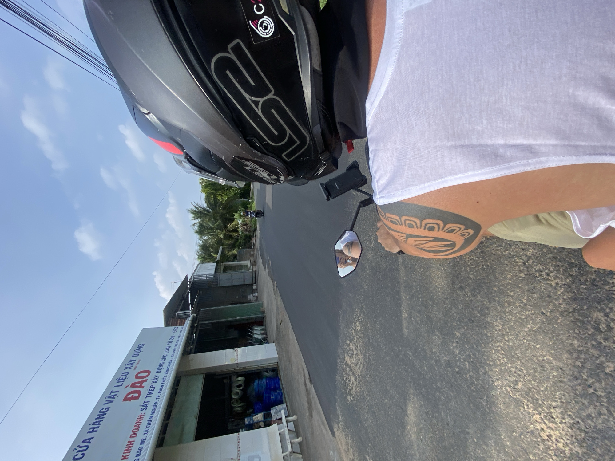 The image shows a person riding a motorcycle in Mũi Né, Vietnam. The person is wearing a black LS2 helmet and a white tank top, with a tribal tattoo visible on their arm. The motorcycle has a phone mount attached to it. In the background, there is a building with a sign that reads "CỬA HÀNG VẬT LIỆU XÂY DỰNG ĐÀO". The building appears to be a construction materials store, with various items visible inside, including blue barrels and what look like pipes. The road is asphalt, and there are palm trees lining the sides. The sky is blue with some white clouds. Another person on a motorcycle can be seen further down the road. The scene is illuminated by sunlight.