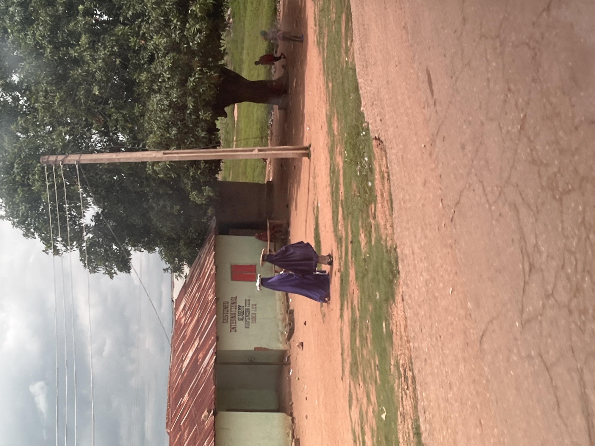 In Karshi, Nigeria, a light green building with a corrugated iron roof, labeled "Destined International Academy," stands on a dirt road. Two figures in dark purple and black robes walk away from the building. A large tree stands to the right of the building. In the background, a red-shirted child runs, and another figure stands near the tree. The sky is overcast.