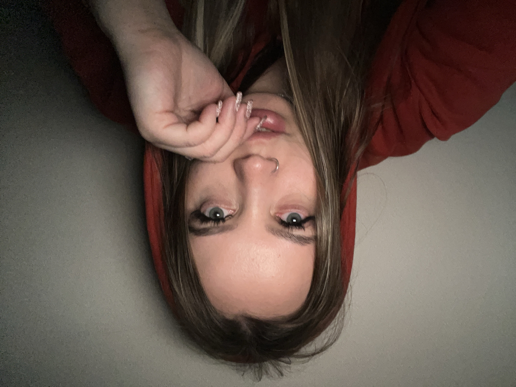 A close-up, inverted portrait features a young individual, likely female, with long brown hair cascading around her face, which appears to be styled with lighter streaks. Her head is positioned upside down in the frame. She has striking blue eyes, enhanced with prominent dark eyeliner and mascara, looking directly towards the viewer. Her lips are full and slightly parted, and she wears a silver septum piercing in her nose. Her right hand, with long, glittery light-colored fingernails, is brought up near her mouth, with fingers gently touching her lower lip and chin area. She is wearing a red hooded garment, possibly a hoodie, which frames her face. The background consists of a plain, light grey wall, indicating an indoor setting. The image captures a casual, intimate moment, likely a selfie. No specific time of day, weather conditions, or textual elements are discernible.