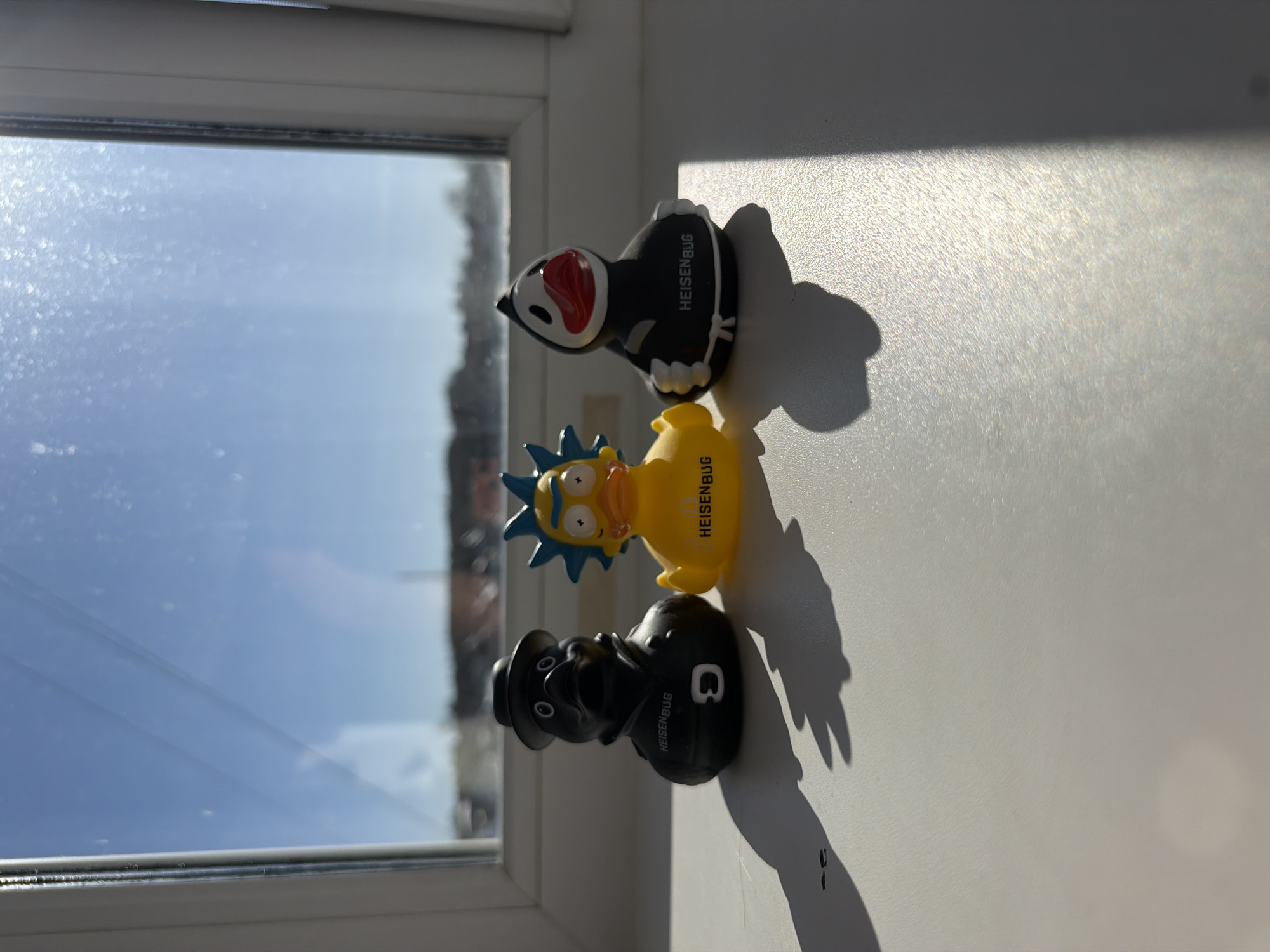 Three distinct collectible figures, styled like rubber ducks, are arranged in a line on a white windowsill. From back to front, the figures include a black and white character with a prominent red beak-like mouth, a yellow figure with blue spiked hair and large eyes reminiscent of Rick Sanchez from *Rick and Morty*, and a black figure wearing a fedora, strongly resembling Walter White (Heisenberg) from *Breaking Bad*. Each of the three figures is inscribed with the text "HEISENBUG." A partial letter, possibly a "C" or "Q," is also visible on the back of the black fedora-wearing figure.
The figures cast long, dark shadows onto the textured white surface of the windowsill, indicating strong, direct sunlight. To their left, a window reveals a clear blue sky, suggesting bright and sunny weather conditions. Some dust or minor imperfections are visible on the windowpane. The scene is illuminated by natural light, pointing to a daytime setting, likely morning or late afternoon given the angle and length of the shadows. The image captures a quiet indoor scene in Chaikovsky, Russia.