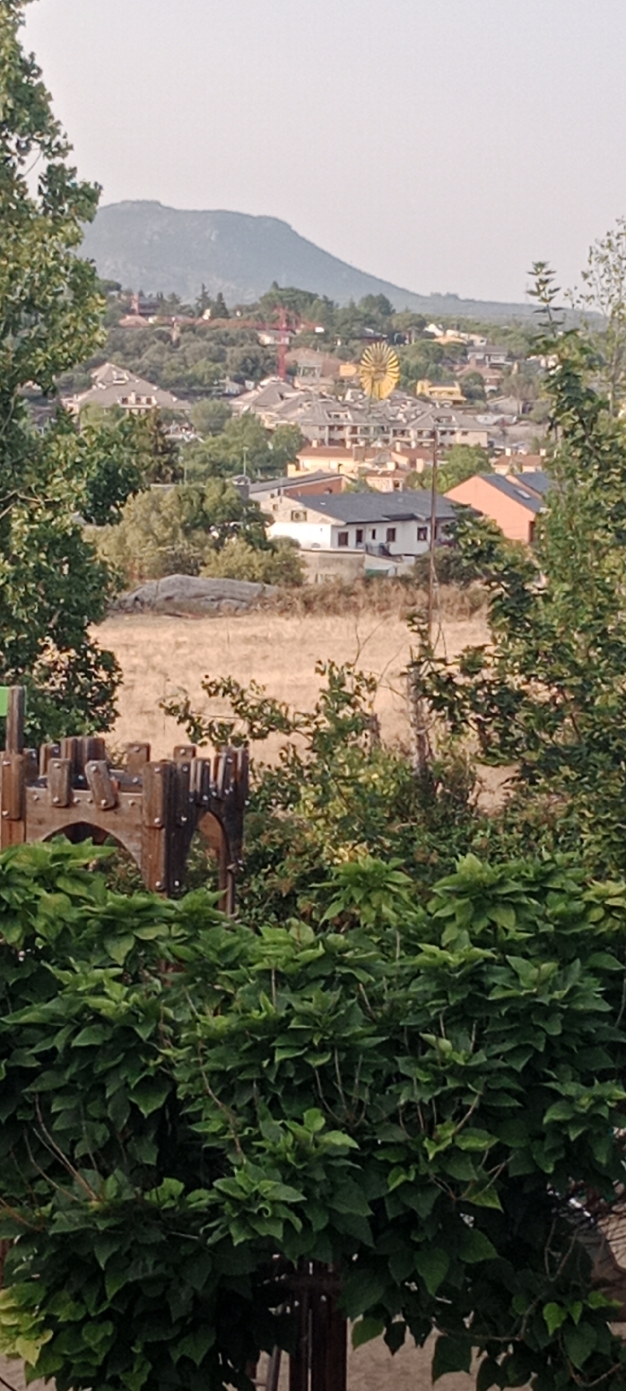 This is a vertical, medium shot photograph taken from a slightly elevated perspective, looking out over a town nestled in a valley with a large hill in the background. The foreground is dominated by lush green foliage, obscuring the lower part of the scene. To the left and slightly behind the foliage, a wooden playground structure resembling a castle turret with crenellations and arched openings is visible.
Beyond the foliage and playground, a dry, grassy field stretches out, dotted with scattered rocks. Further in the mid-ground, a cluster of buildings forms a town. The architecture appears to be primarily residential, with roofs of various colors including grey, orange, and white. Several houses feature dark-tiled roofs, and some have visible windows and balconies. A prominent yellow, sun-shaped structure, possibly a decorative windmill or a piece of public art, stands out among the buildings. A red construction crane is also visible in the distance.
The background features a large, rounded hill that dominates the horizon. The sky is overcast and appears hazy or foggy, obscuring the very top of the hill and the far distance. The overall scene suggests a suburban or semi-rural environment in Spain, likely during daylight hours with a muted light due to the overcast conditions. Based on visual cues and location information, this image is likely of the town of Galapagar, Spain, given the distinctive topography and architectural style common in the area. There are no people or animals visible in the photograph.