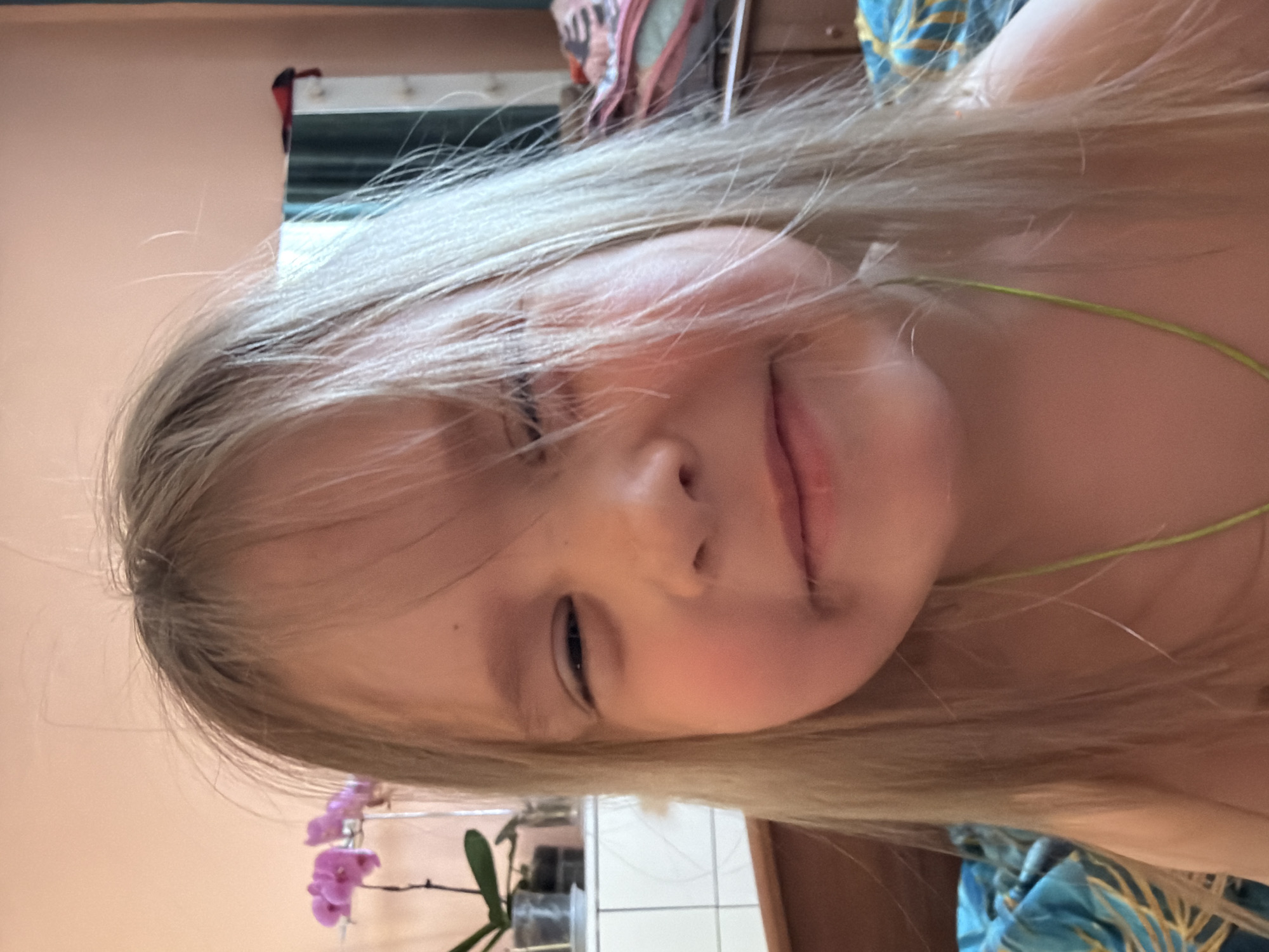 A close-up portrait of a young girl with blonde hair is seen with her head tilted to the right. The child has light skin, a small nose, and a slightly closed left eye. Part of a light green necklace is visible at the right of the frame. Behind her, there are elements of an interior setting, including a partially visible mirror and furniture. In the bottom left corner, a pink orchid is in focus. Based on the visual context, this image might have been taken indoors. There's not enough information from the image to determine a location in the city of Chaikovsky, Russia.
