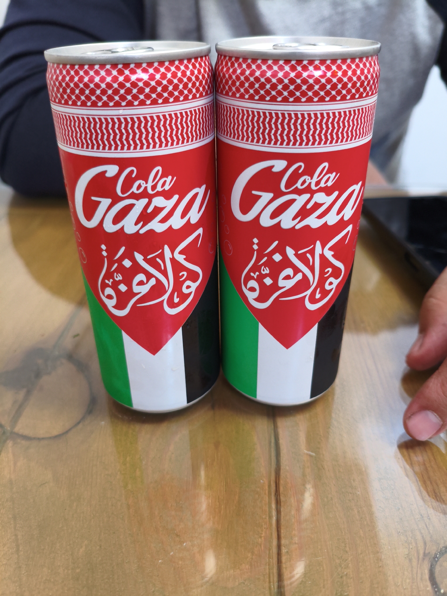 Two cans of "Gaza Cola" are displayed on a wooden table. The cans have a red base with white lettering that reads "Cola Gaza" and Arabic script below. The cans are decorated with a pattern resembling a Keffiyeh and the Palestinian flag colors (green, white, and black) at the bottom. The table surface is reflective and shows wood grain. In the background, a person's torso wearing a dark grey shirt is visible. To the right of the cans, a hand with visible fingernails is reaching towards the right edge of the frame. The scene is likely indoors, with the lighting suggesting a moderately lit environment. This scene is not located in Cambridge, United Kingdom.