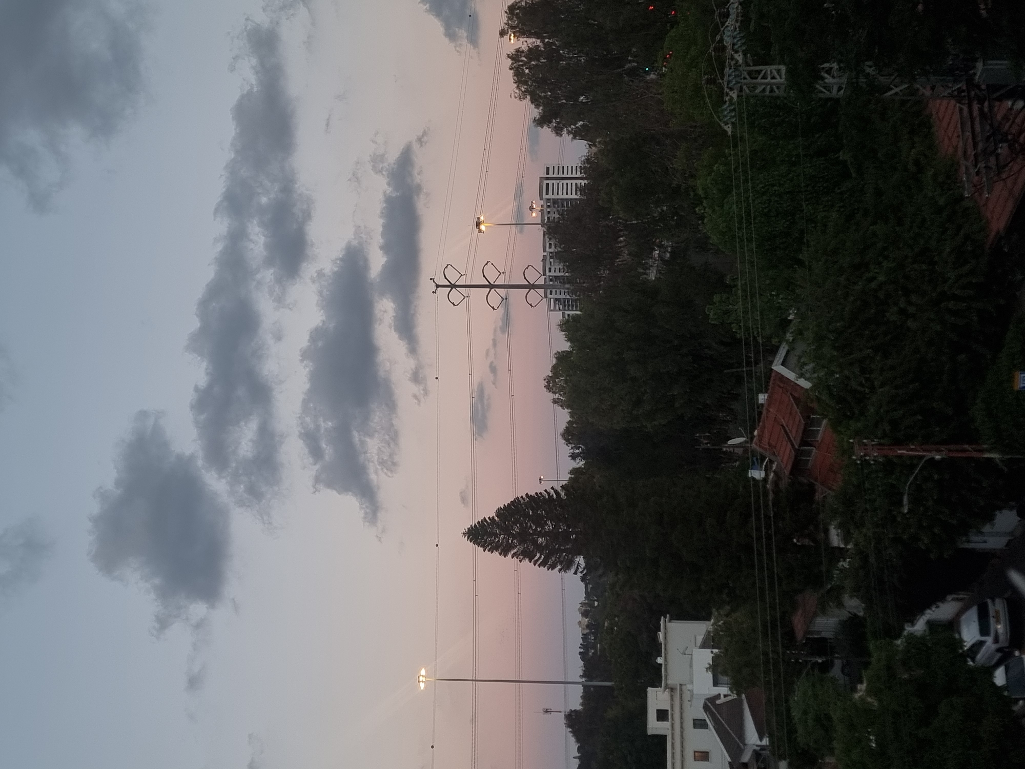 The image shows a cityscape at dusk in Ramat Gan, Israel. The sky is partially cloudy, with dark gray clouds contrasting against a pinkish-gray hue. In the midground, electrical pylons and wires stretch across the scene. Streetlights illuminate the area. Buildings are interspersed with trees. Houses with red-tiled roofs and white apartment buildings are visible. Cars are parked along a road.