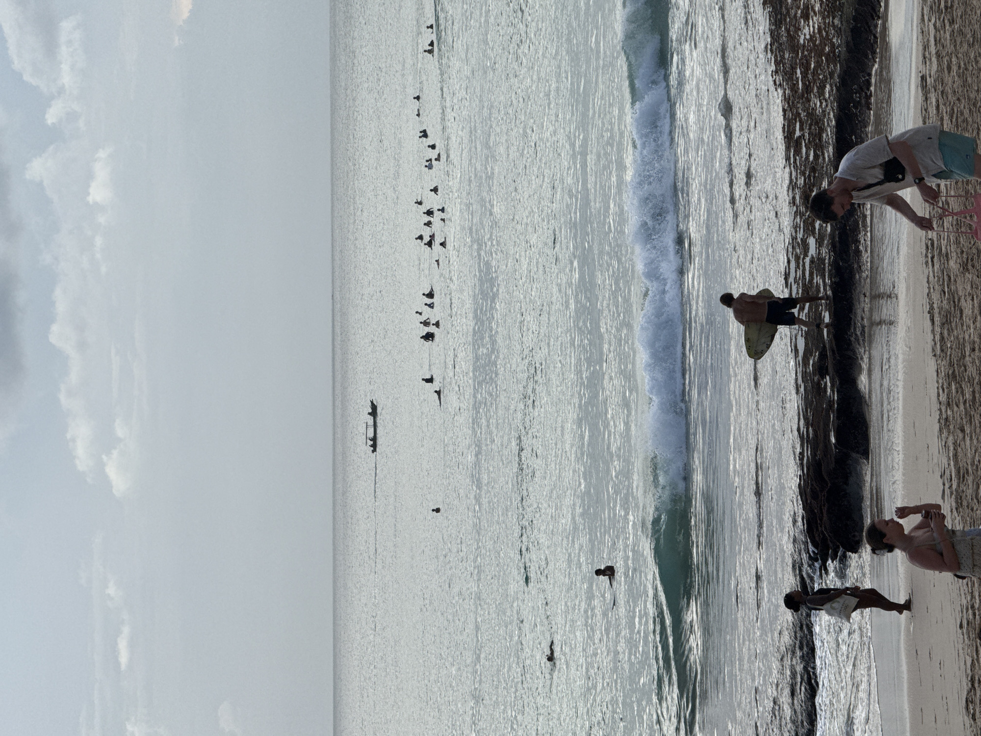 This image captures a lively beach scene, likely in Kuta Selatan, Indonesia, with a vast expanse of ocean under a hazy or overcast sky. The overall lighting is bright but diffused, creating a silvery, reflective surface on the water.
In the foreground, the sandy beach features darker, possibly wet or rocky patches at the water's edge. Several individuals are present on the shore. One man, wearing a light t-shirt and light blue shorts, bends down, holding a small pink object. Nearby, another person stands partially visible. Further along the beach, a woman in a light top looks towards the water, and another person in darker attire stands closer to the incoming waves.
In the shallow water near the shore, a man stands with a yellow surfboard, seemingly preparing to surf or having just exited the water. A significant group of approximately 20-30 people are scattered further out in the ocean, mostly silhouetted against the bright water. They appear to be surfers or bodyboarders, patiently waiting for waves to break. Their postures suggest they are on or near boards, ready to ride. A few other individuals can be seen swimming or floating closer to the shore.
Even further in the distance, a small, elongated boat with a few occupants is visible on the calm horizon, suggesting fishing or transport activities. The scene conveys a relaxed, recreational atmosphere typical of a popular surfing beach. No visible text is present in the image.