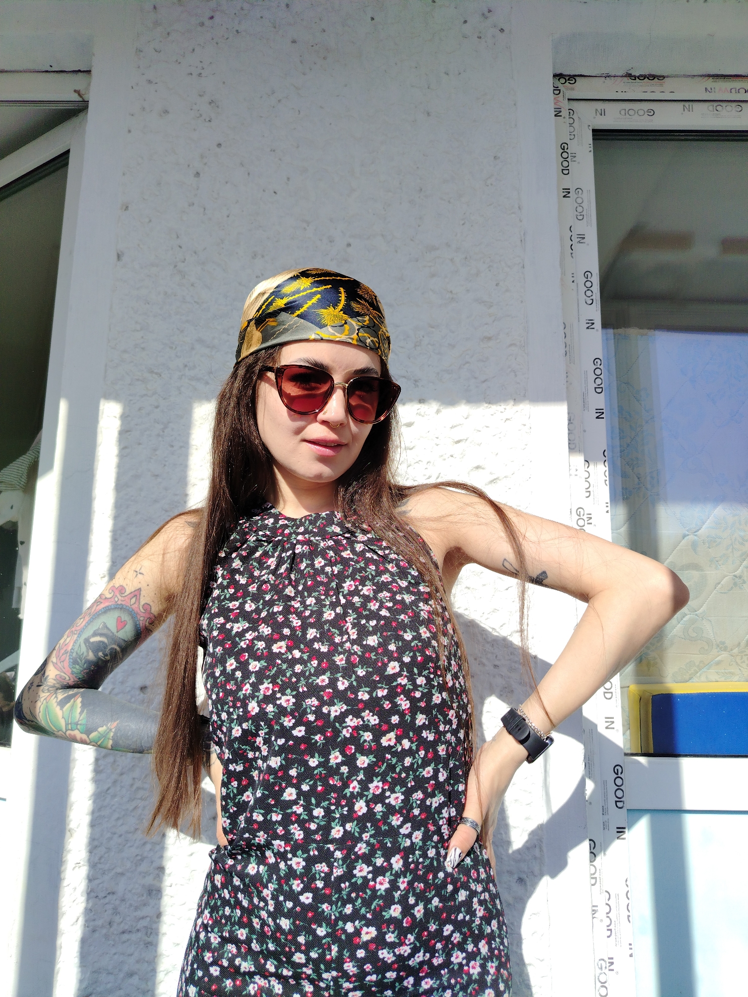 The image shows a woman with long brown hair, wearing a headscarf and sunglasses, standing in front of a white wall with a window. She has tattoos on her arms and is wearing a floral print dress and a watch on her left wrist. She has her hands on her hips. The window frame has the words "GOOD IN" printed on it repeatedly. The location is the city of Noviy, Russia.