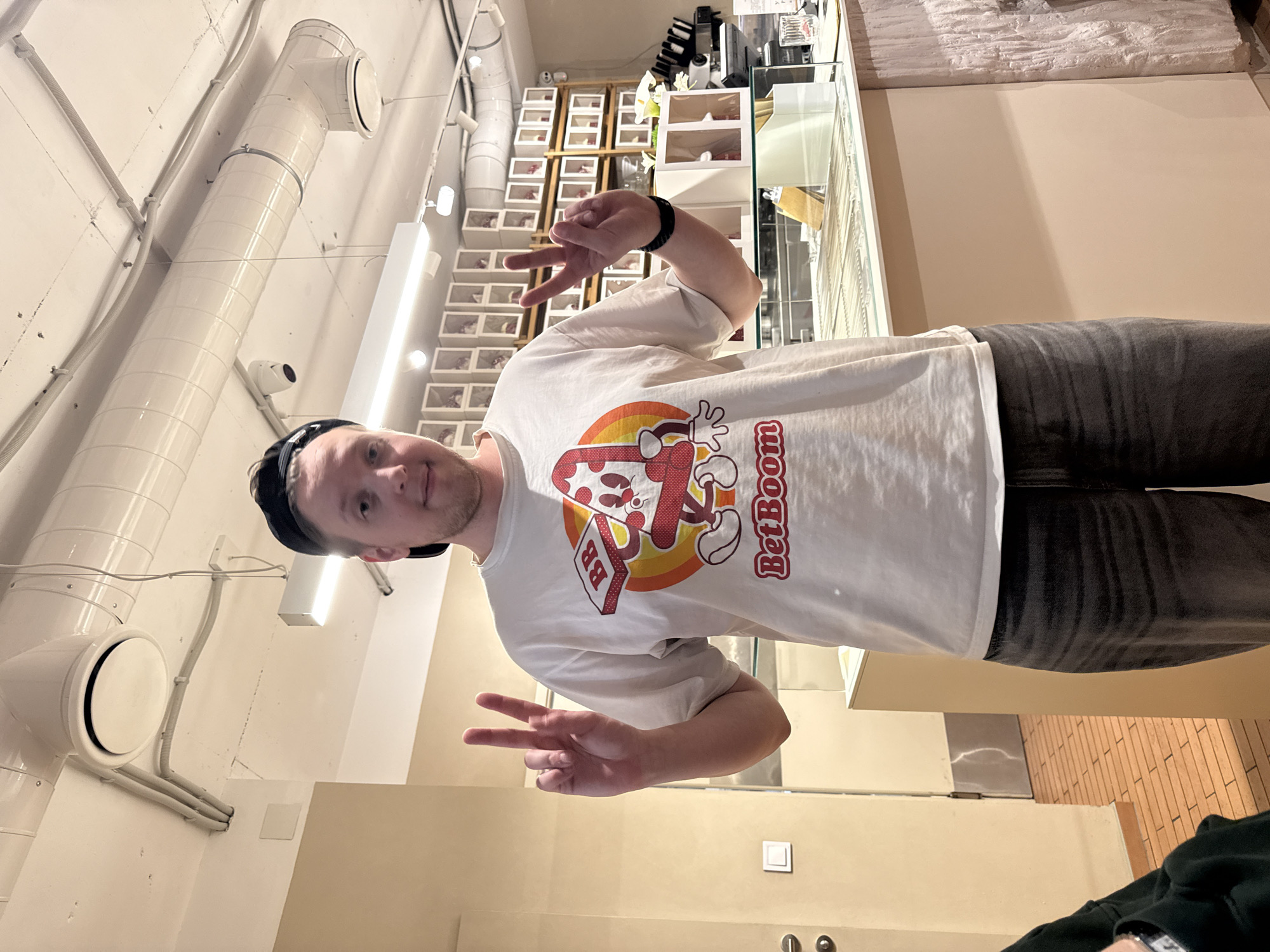 A Caucasian male, fair-skinned, with short blonde hair and stubble, is positioned indoors. He wears a dark baseball cap and a white t-shirt with the "BetBoom" logo. He is wearing a wristband. He is oriented towards the camera. He is making the peace sign with both hands.
The backdrop suggests a retail or commercial space, with rows of boxes displayed on shelves. Fluorescent lighting and industrial-style air ducts are prominent overhead. It is likely in Belgrade, Serbia.