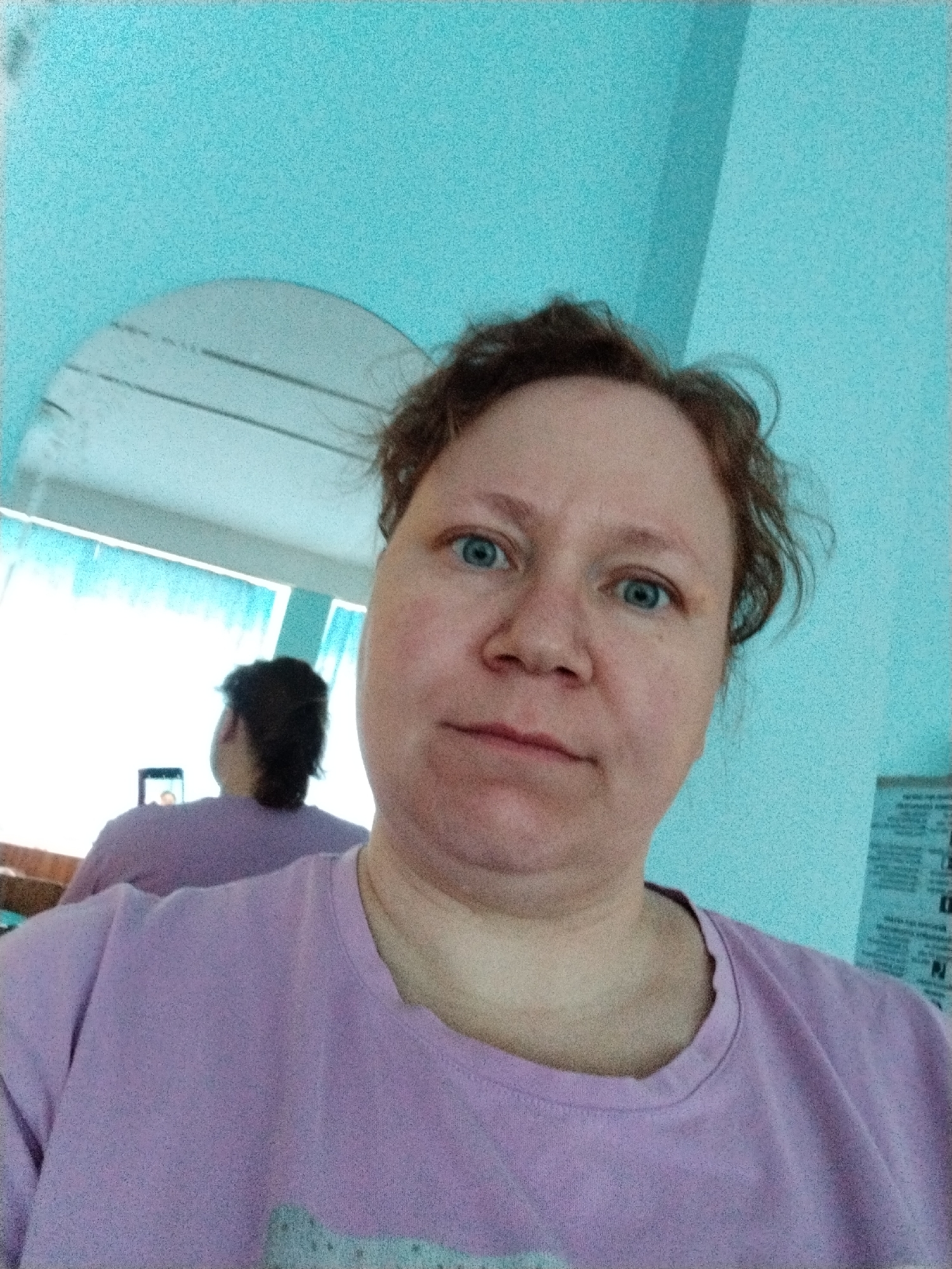 A woman with light skin, blue eyes, and reddish-brown, curly hair takes a selfie, looking directly at the camera with a neutral to slightly pensive expression. She is wearing a light purple (lavender) t-shirt.
Behind her, her reflection is visible in a semi-circular mirror, showing her from behind, also wearing the same purple t-shirt. The reflection reveals a bright window behind her, suggesting daylight. In the reflection, she appears to be holding or looking at a dark-colored smartphone or device.
The immediate background is dominated by a vibrant turquoise or light blue wall, which is visible above and to the sides of the woman. On the far right, a white sheet of paper with indistinct black text, possibly a document or poster, is partially visible against the blue wall. The overall image has a grainy texture. The scene takes place indoors, in Chaikovsky, Russia.