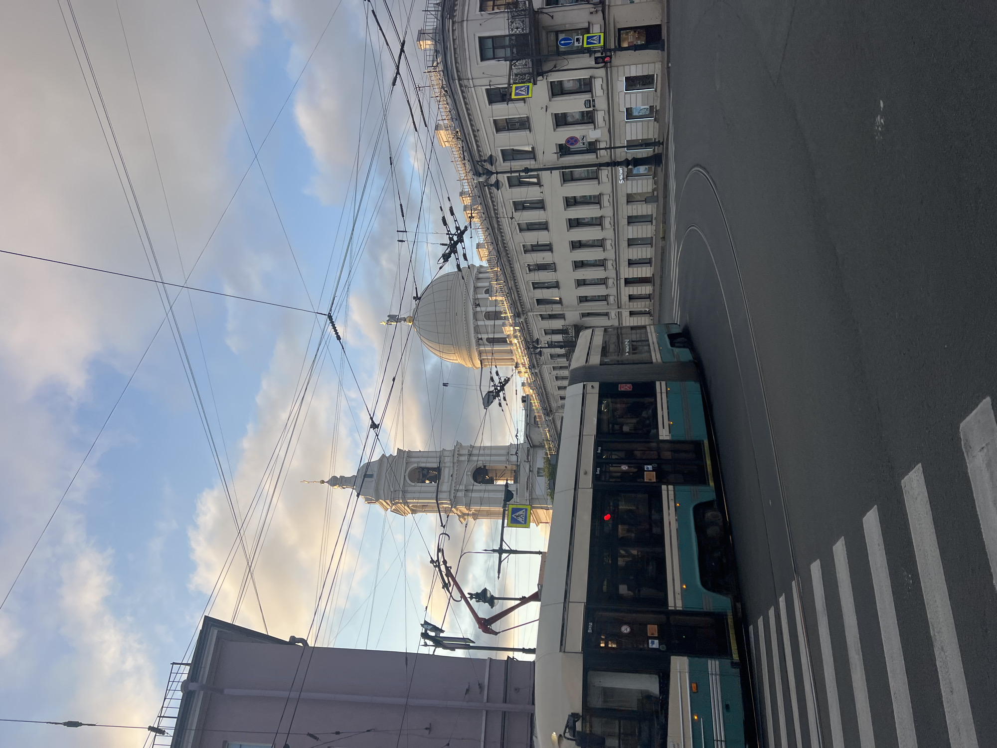 The image depicts a street scene in Saint Petersburg, Russia, at the intersection of Nevsky Prospekt and Sadovaya Street. A modern tram, with a cream-colored top and teal lower body, is turning right, its pantograph extended and connected to overhead wires. The tram has "ВХОД" (entrance) visible on its side. In the background, a large white building with a dome, likely the Kazan Cathedral, is prominent. To the left of the dome, a tall, white bell tower, also part of a religious structure, rises above other buildings. The sky is partly cloudy, with the sun casting a warm light on the scene. The street is asphalt, with a white pedestrian crossing marked on the left side of the image. Traffic lights and street signs are visible, indicating a regulated intersection. The buildings are multi-story, featuring classical architectural styles with numerous windows. No people are clearly visible in the image. The overall impression is of a busy urban environment in Saint Petersburg during daylight.