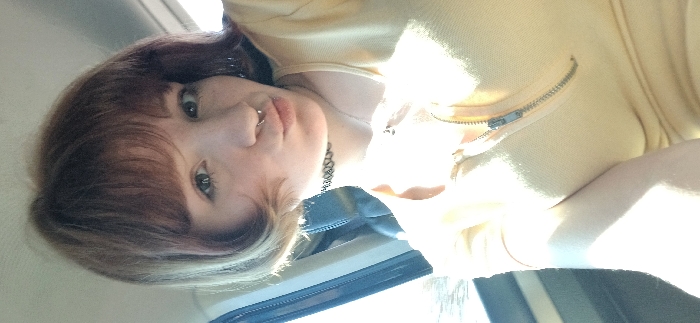 A single individual, identified as a female, is depicted in a close-up, front-facing perspective, likely a selfie. She has reddish-brown, chin-length hair with lighter blonde or light brown tips, styled with bangs across her forehead. Her eyes are light-colored, and she has visible freckles on her face. A silver-toned septum piercing is visible in her nose, and her lips are puckered. She wears a black choker necklace and a silver-colored chain necklace with a small, dark pendant. Her attire consists of a light yellow short-sleeved top with a metallic zipper running vertically down the front, partially unzipped.
The setting appears to be the interior of a vehicle, possibly in the front passenger seat. A dark, upholstered seat or seatback is visible behind her left shoulder. To the left of the frame, a window frame and bright outdoor light are apparent, causing significant overexposure in that area and casting strong illumination on the subject's left side. The upper part of the background, above her head, is a light, indistinct surface, possibly the vehicle's ceiling. The image provides no specific visual cues to confirm the stated location of Bistrice, Montenegro.