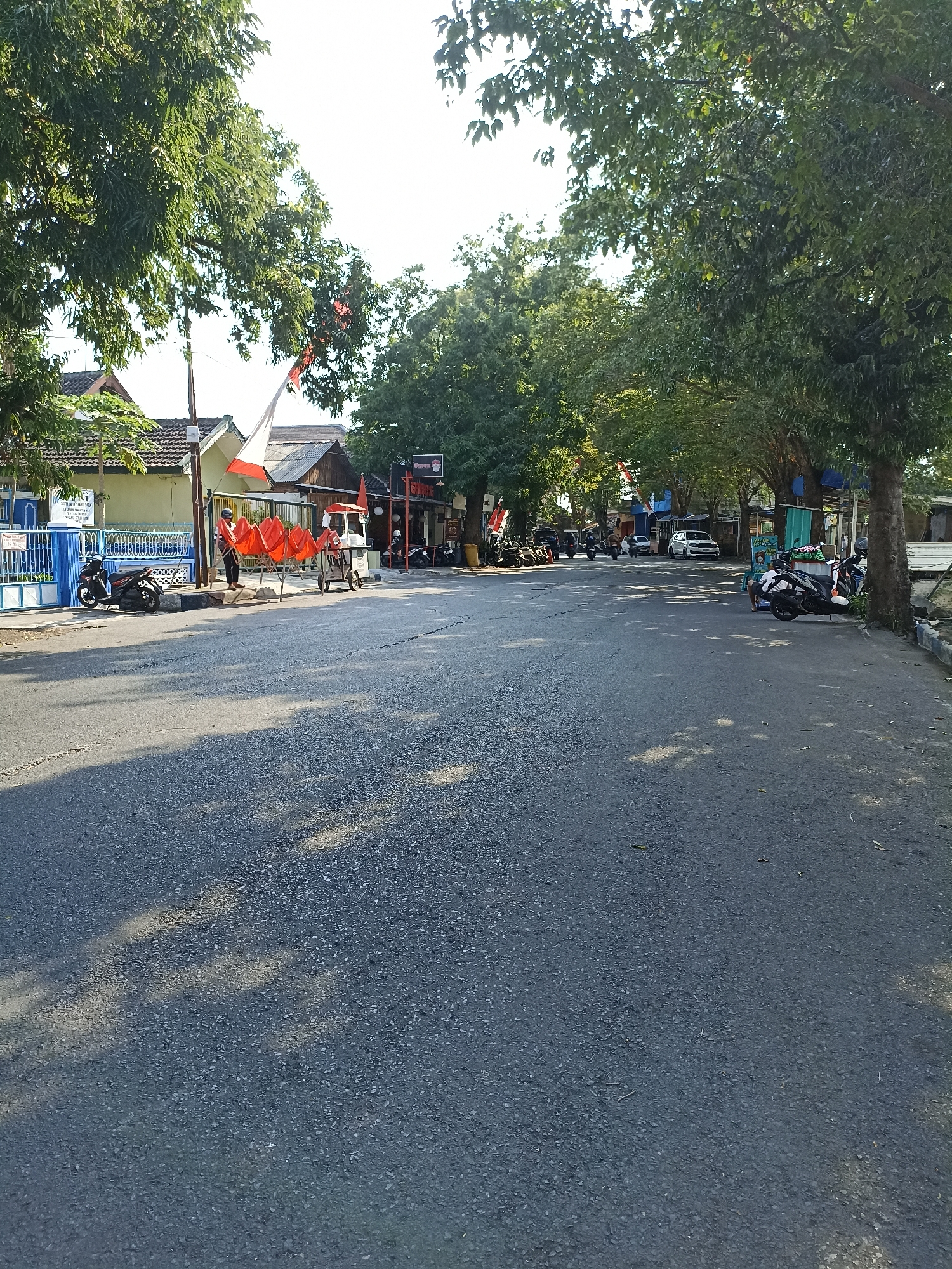 The image depicts a street scene in Lumajang, Indonesia, under a bright sky with dappled sunlight filtering through overhead trees. On the left side of the street, a blue gate with white trim is visible in front of a building with a tiled roof. A black motorcycle is parked near the gate. Further along the street on the left, several orange seats are set up on a metal frame, with a person standing nearby, wearing a helmet. A small food cart is also present. Across the street, several more motorcycles are parked. A white car is driving in the distance, and further down the road, there appear to be shops or stalls with awnings. The road surface is asphalt, and trees line both sides of the street, creating a canopy effect. Indonesian flags are displayed on poles in front of some buildings.