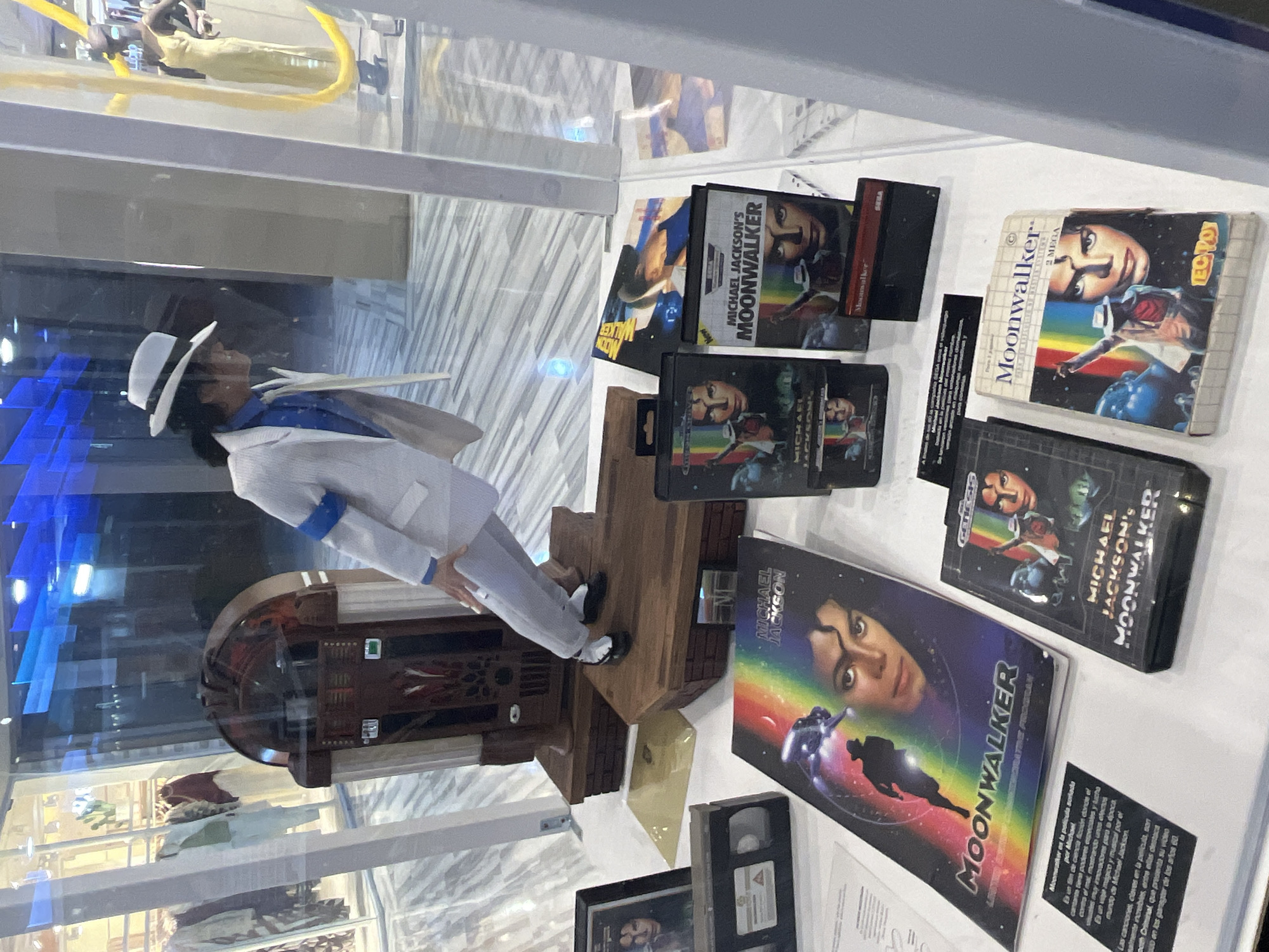 This image captures a display of Michael Jackson memorabilia, likely within a museum or exhibition in Las Palmas, Spain. The main subject is a detailed figurine of Michael Jackson, dressed in a white fedora, white suit with a blue armband, black shirt, and black shoes, performing his iconic anti-gravity lean. He stands on a wooden base that incorporates a miniature replica of a jukebox, reminiscent of the "Smooth Criminal" music video set.
Arranged on the white surface around the figurine are various "Moonwalker" video games and media. These include multiple game cartridges and boxes for "Michael Jackson's Moonwalker" across different platforms such as SEGA Genesis and Mega Drive, as indicated by labels like "GENESIS," "SEGA," and "2 MEGA." There is also a larger video cassette or laserdisc cover for "Moonwalker," featuring a prominent image of Michael Jackson's face and a silhouette of him dancing in space with a rainbow streak.
A plaque next to the media provides text in Spanish. It begins with "MOONWALKER" and describes the film as Michael Jackson's "dreamed film," a science fiction movie where the singer has special powers and fights evil, showcasing innovative visual effects and a magical musical journey. It highlights the incredible songs, particularly "Smooth Criminal," which features a music video set among 1960s gangsters. The objects are displayed under glass, reflecting the ambient lighting of the indoor venue.