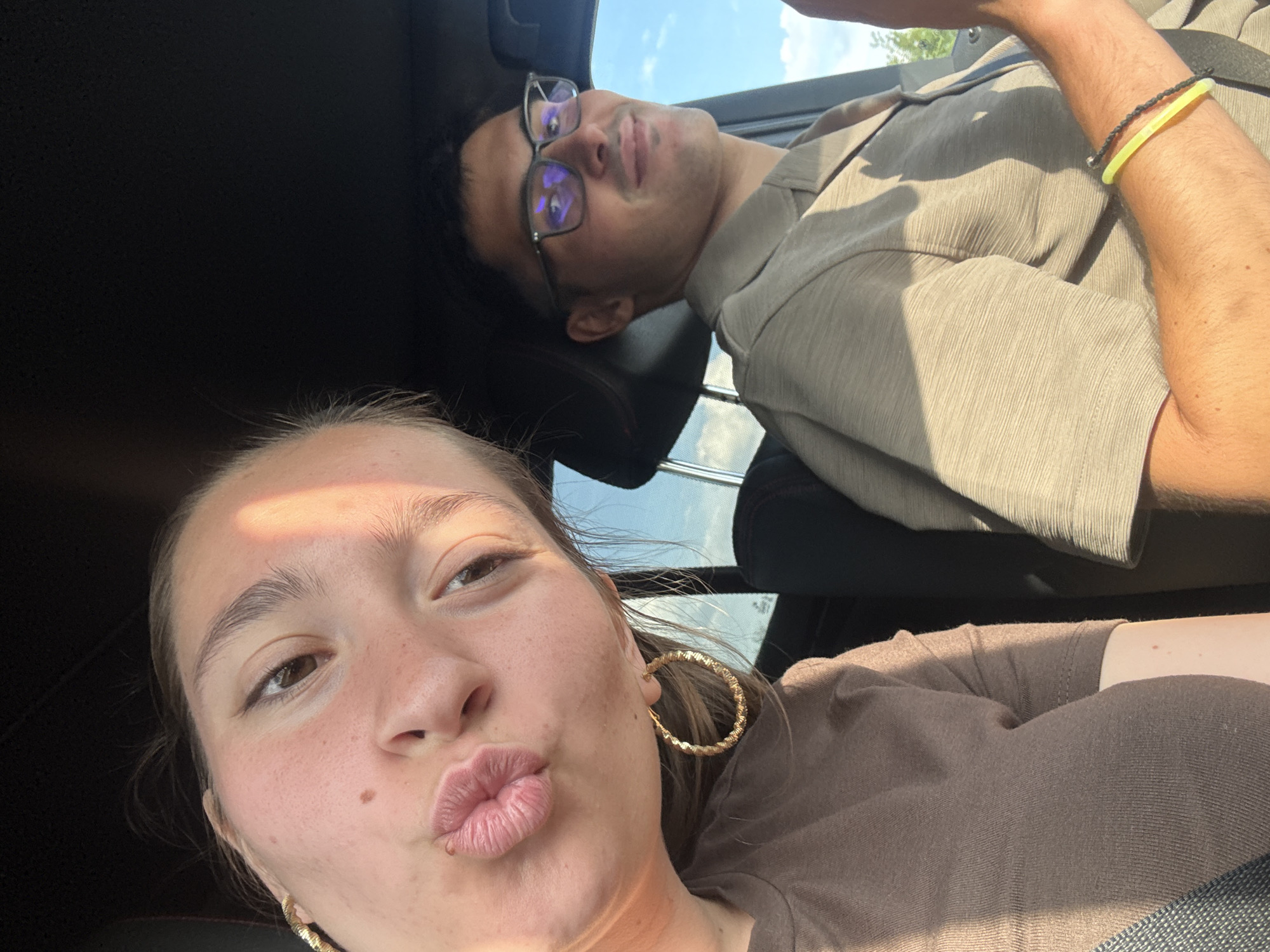 The image shows a selfie taken inside a vehicle, likely a car. Two individuals are visible, a woman in the foreground and a man in the background. The woman, positioned on the left, leans in towards the camera with her face angled to the side, puckering her lips. She has fair skin, brown eyes, and is wearing a brown t-shirt and large gold hoop earrings. Sunlight casts a warm glow on her face. The man, on the right, is wearing glasses and a light-colored collared shirt. He is looking towards the camera with a slight smile. His arm is visible, and he is wearing a dark bracelet with a neon yellow element. The interior of the car is dimly lit, with the dashboard and seats suggesting a typical vehicle interior. The background outside the car window shows a clear blue sky with some greenery. The scene appears to be a casual moment captured while traveling. The mention of Rupkite, Bulgaria, in the prompt does not correlate with any visual information present in the image.