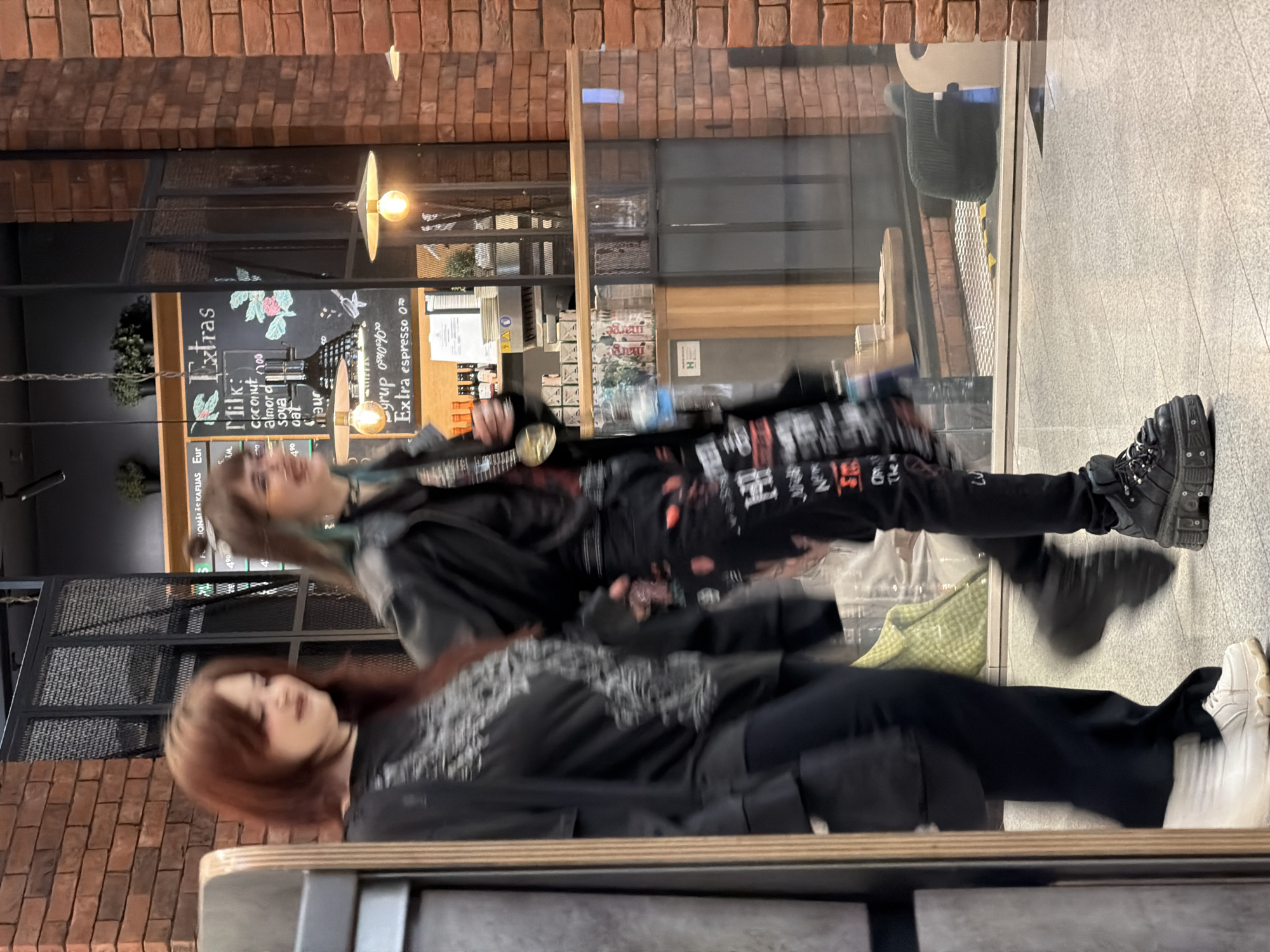 Two women stand in front of a storefront in Riga, Latvia. The building's exterior features a brick facade. The storefront's window displays a menu board listing coffee extras and milk options, including coconut, almond, soy, and oat milk. The women are dressed in dark clothing. The first woman's outfit features a graphic design. Both are wearing black pants. The second is also wearing white shoes. The location appears to be a coffee shop.