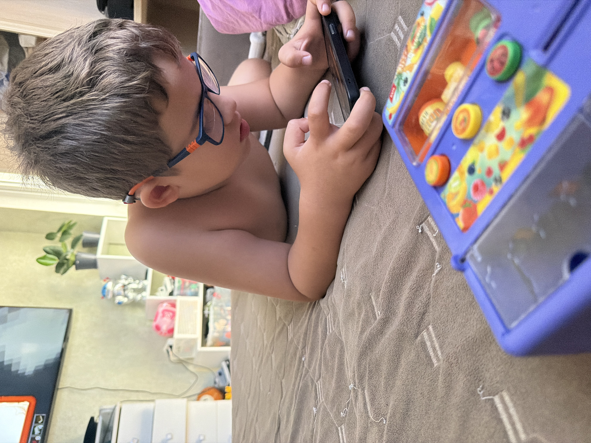 A young child, likely a boy, lies shirtless on his stomach on a textured brown blanket, fully engrossed in using a smartphone. He wears blue and orange glasses and holds the black-cased phone with both hands, his eyes fixed on the screen. Beside him, to the right, is a bright purple toy, possibly a play refrigerator or interactive unit, decorated with colorful fruit illustrations and featuring several round, brightly colored knobs (green, yellow, orange). The toy is not actively being played with by the child.
The setting is indoors, characteristic of a casual home environment in Sochi, Russia. In the background, a light-colored wall is visible, along with a small green potted plant and white shelving units holding various objects, some appearing to be toys. A television screen, displaying pixelated graphics that resemble a video game, can be seen on the left side of the frame. The child appears deeply focused on the content on his phone. No legible text is discernible on any of the objects, including the toy or the phone screen.