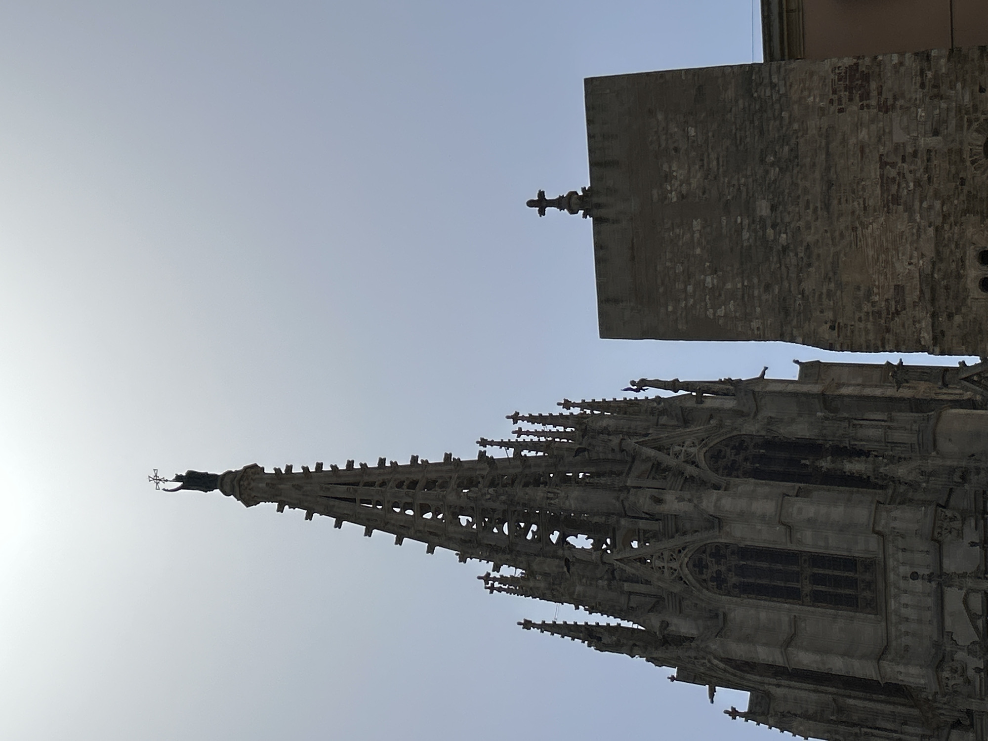 The image shows a low-angle view of two buildings against a bright, hazy sky in Barcelona, Spain. On the left is a tall, ornate Gothic spire with intricate stone carvings and pointed arches. A statue of a figure holding a cross stands at the very peak of the spire. To the right of the spire is a much simpler, square stone tower topped with a cross. The stone of this tower appears older and rougher than that of the spire. The sky is washed out by sunlight, giving the scene a bright but slightly muted appearance. The overall impression is of historic architecture silhouetted against a bright day.