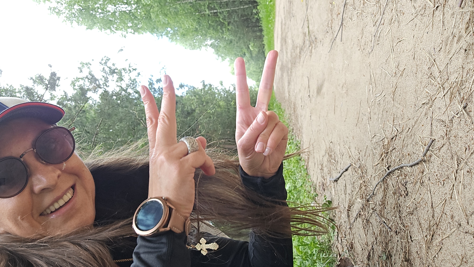 A woman wearing sunglasses and a cap poses for a selfie outdoors in a wooded area. She is wearing a black jacket with a gold cross pendant and a rose gold smartwatch on her left wrist. She is making a peace sign with both of her hands. The background shows a sandy path lined with trees and green foliage. The weather appears to be overcast. This image was taken in Chaikovsky, Russia.
