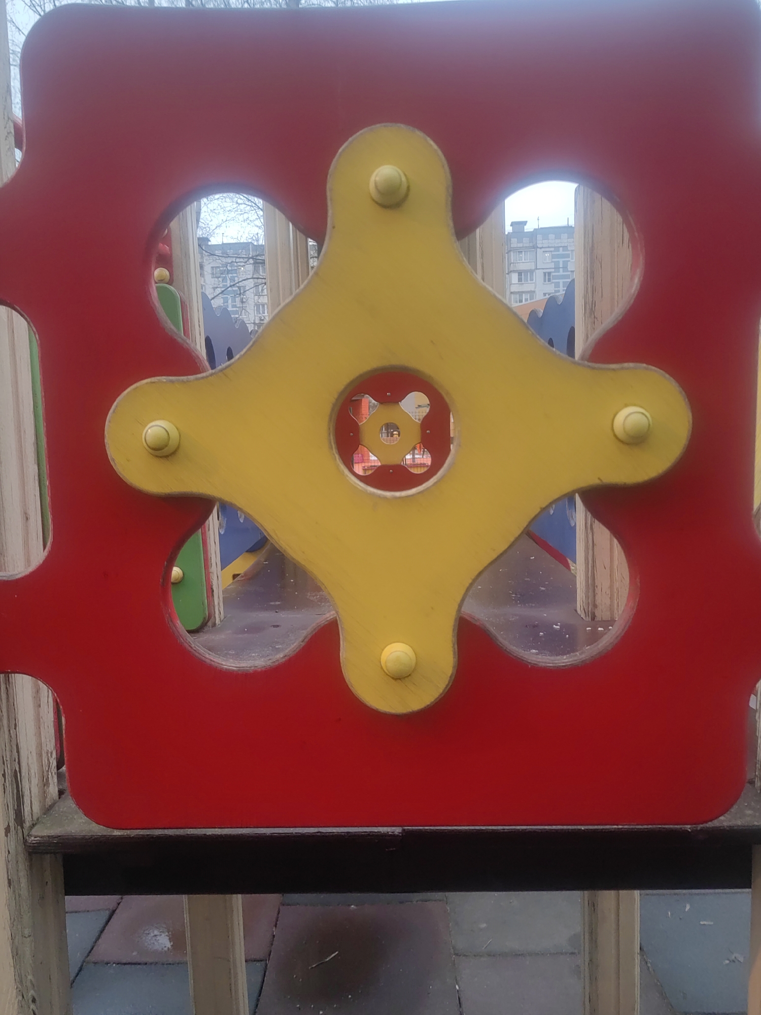 The image captures a close-up view of playground equipment, specifically a section with rotating gears. The main colors are red and yellow. A red panel forms the backdrop, featuring cut-out shapes. A yellow, four-pointed gear is mounted in front of the red panel, secured by round, prominent fasteners. The equipment is positioned on a wooden frame, which is set on a paved surface. The view through the cut-outs shows blurred surroundings, including trees, buildings, and other parts of the playground. The scene suggests an urban playground in Mytishchi, Russia.