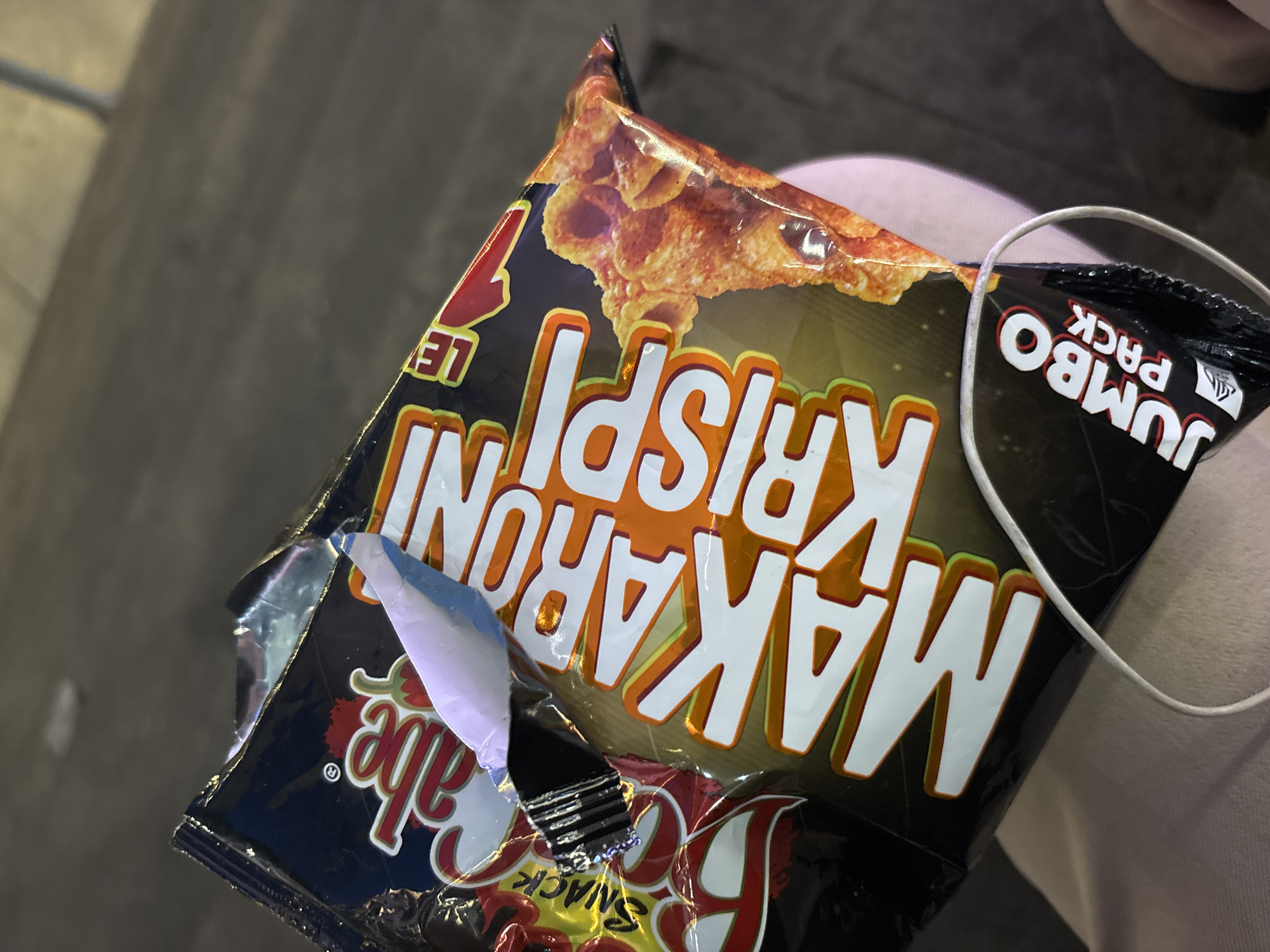 A close-up, slightly angled shot depicts an open bag of "Makaroni Krispi Le" snack chips, specifically a "Jumbo Pack" variant. The bag's packaging is predominantly black and gold with orange and white lettering, featuring an image of the macaroni chips themselves at the top. The bag appears to be torn open, revealing the inside. Part of a white earphone cord is visible against a light tan colored surface, possibly clothing, suggesting the bag is being held. The backdrop is a blurred grey surface. It is highly likely that the image was taken in North Jakarta, Indonesia, as the product is commonly sold there.